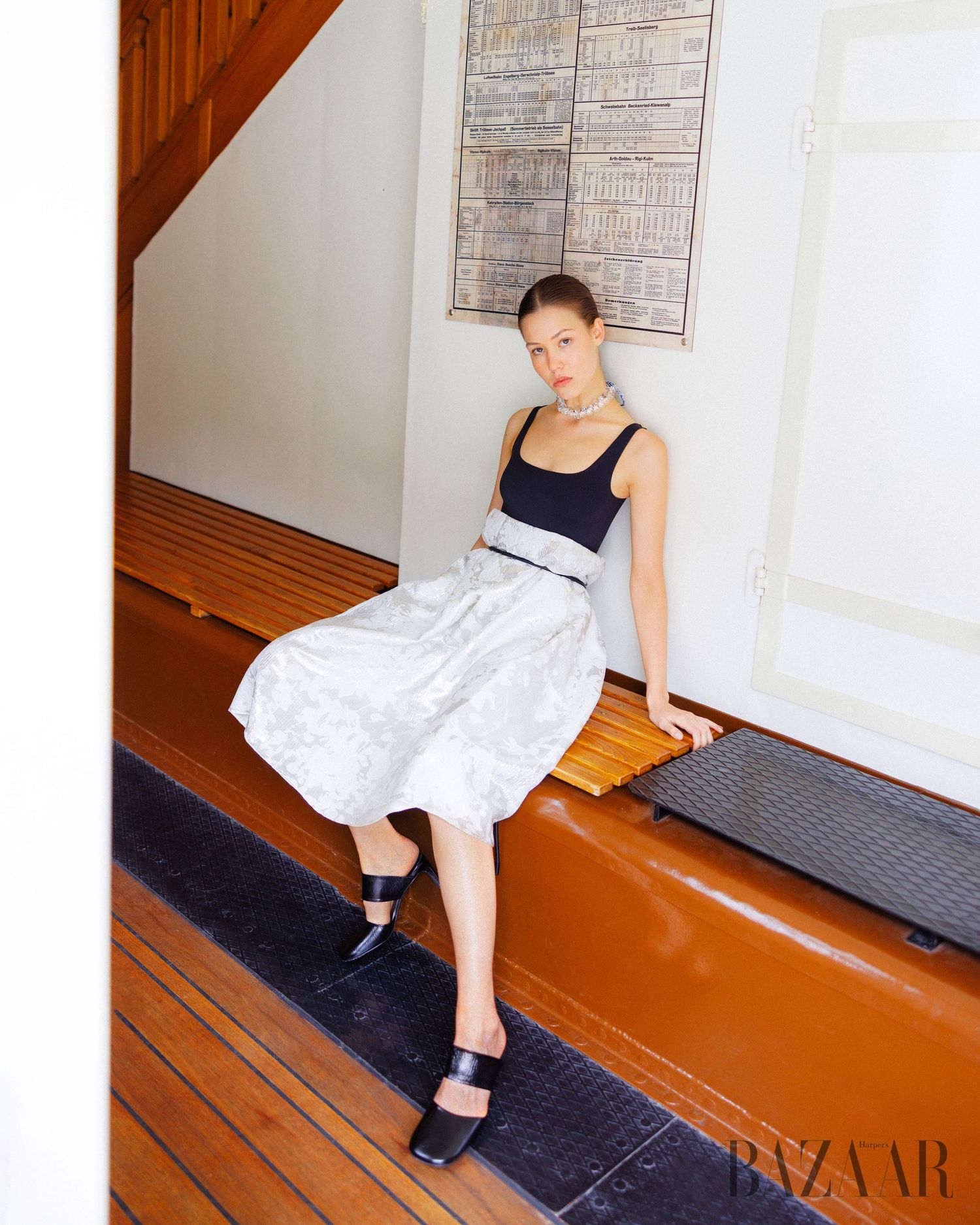 Model in black tank top with silver midi skirt and black mules, seated in a classic interior for Harper's Bazaar.