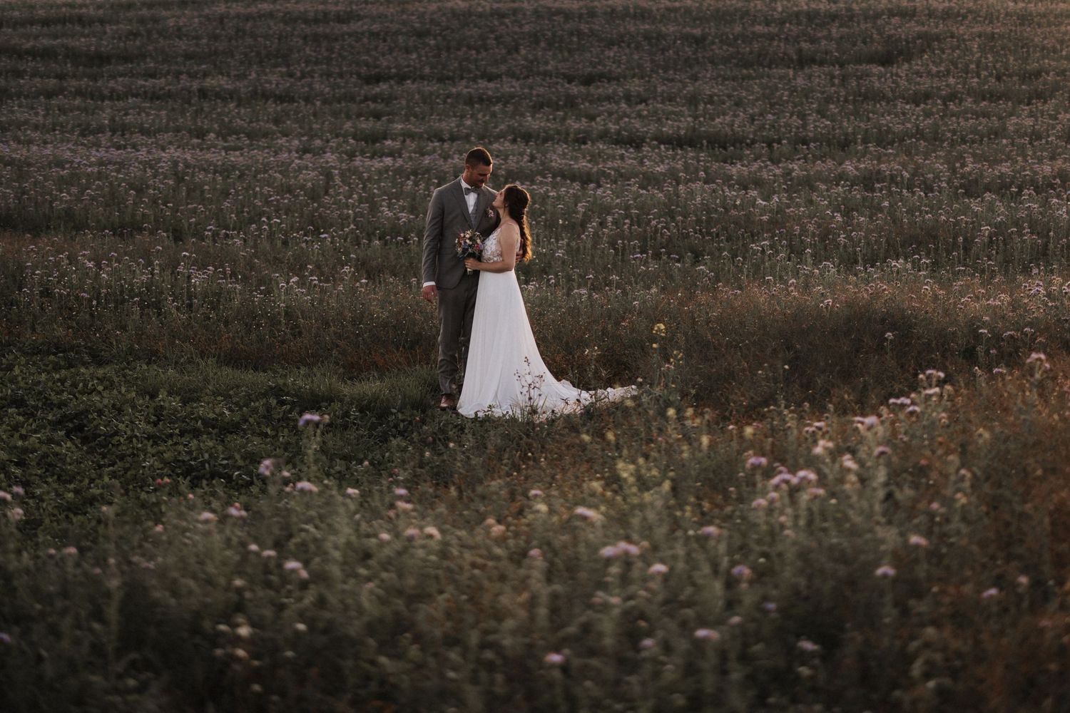 Wedding couple stands together in a wildflower field at dusk, bride in elegant white dress with long train.