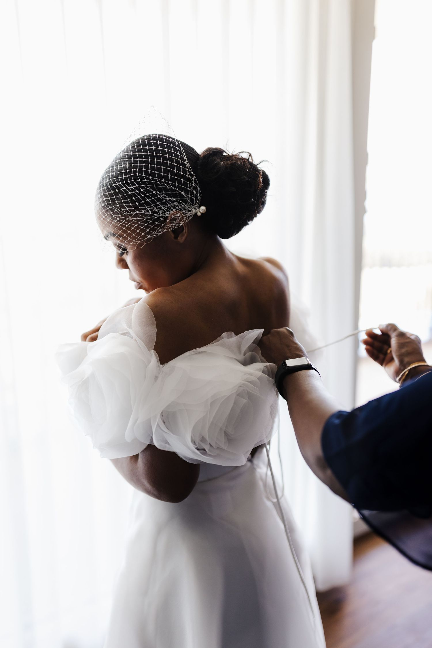 Assistant tying back of white organza wedding dress on Black bride wearing birdcage veil.