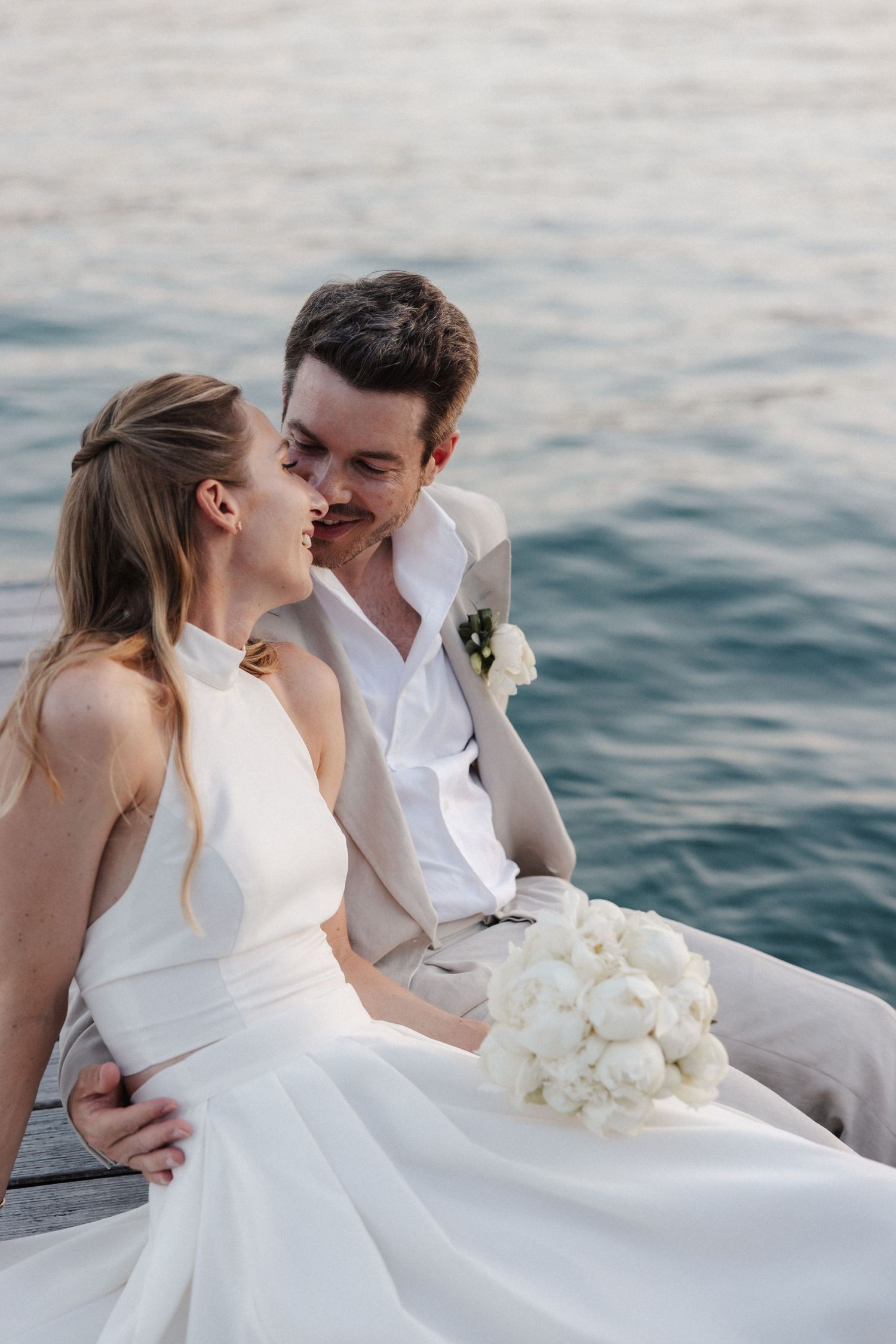 Bride and groom share a romantic moment on a waterfront dock with white bridal bouquet.