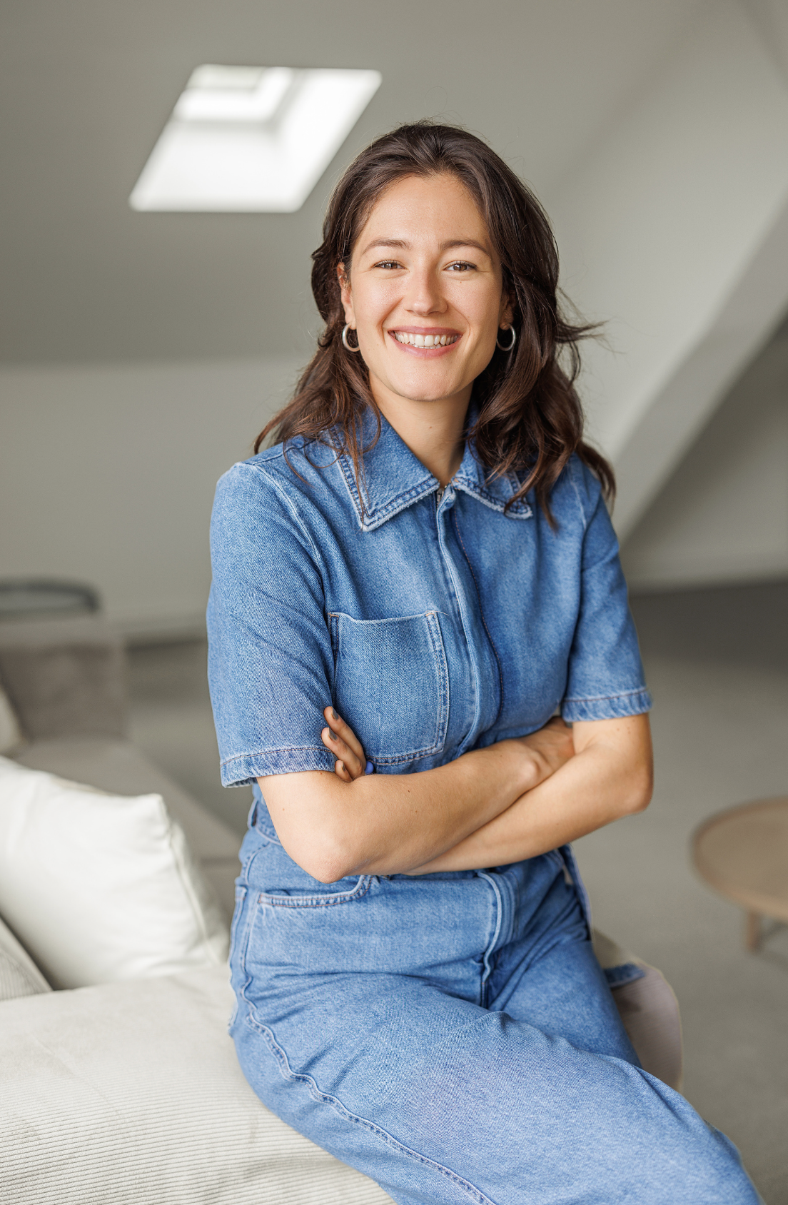 Smiling woman in blue denim dress sitting with arms crossed in bright modern attic room.