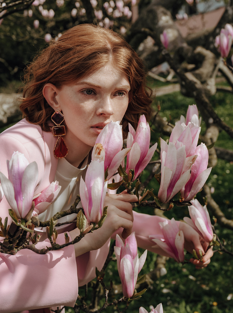 Red-haired woman in pink blazer holding magnolia blossoms on a blooming tree outdoors.