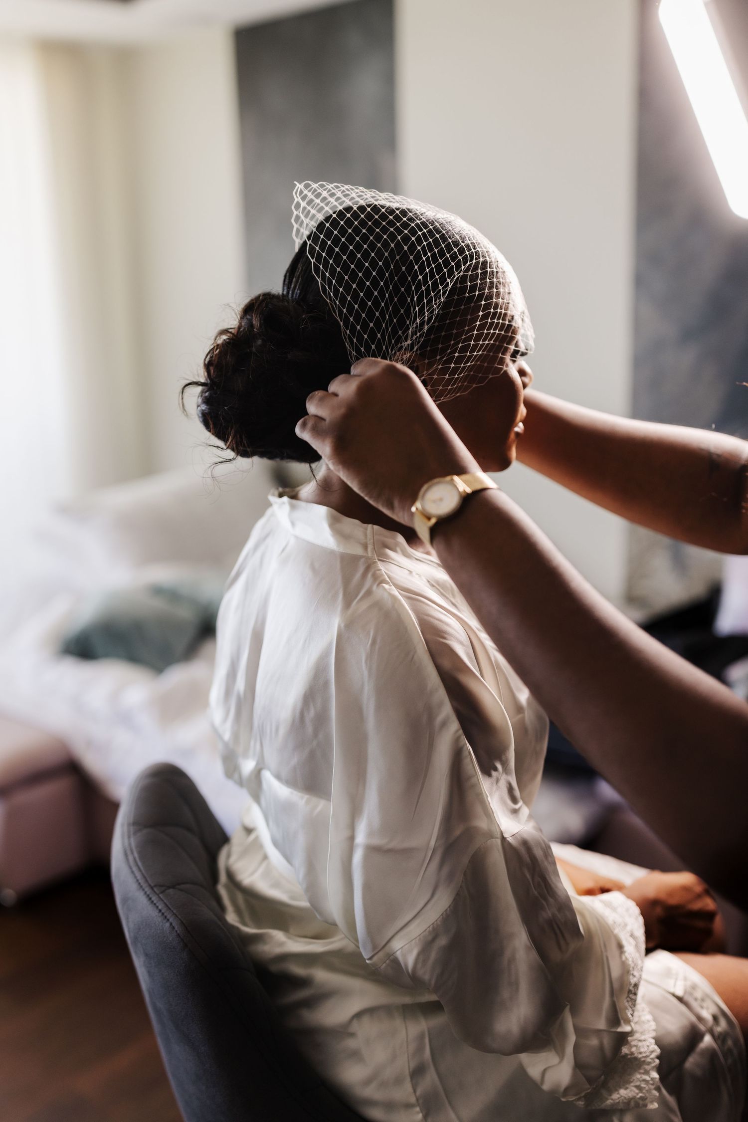 Stylist placing a white birdcage veil on Black bride with updo during wedding morning preparations.