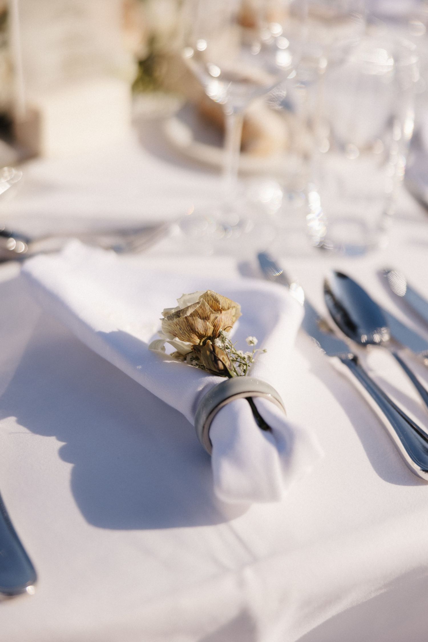 White wedding table setting with rolled napkin adorned with a dried golden rose and silver napkin ring.