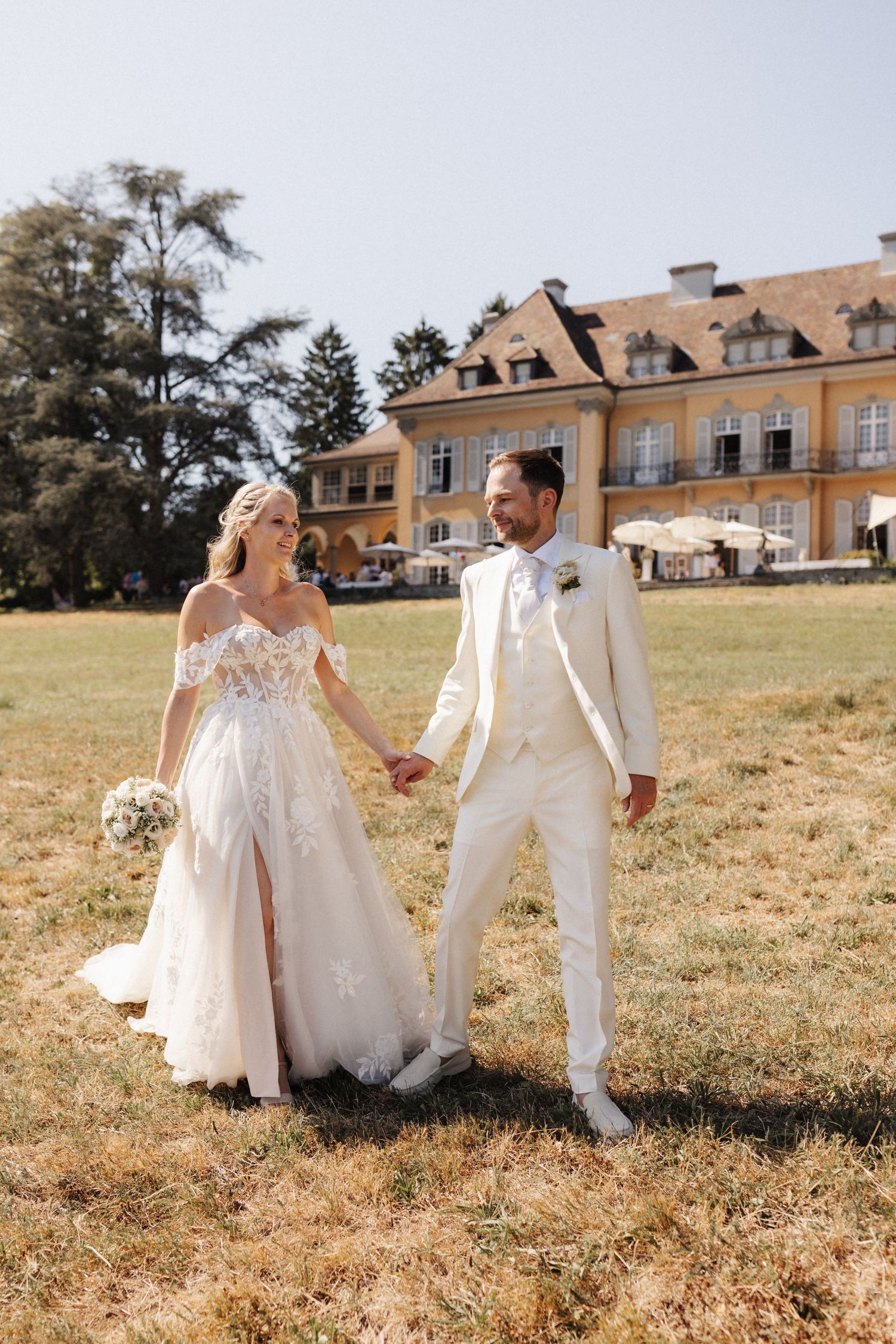 Smiling bride and groom holding hands strolling across a sunny lawn in front of a yellow manor.