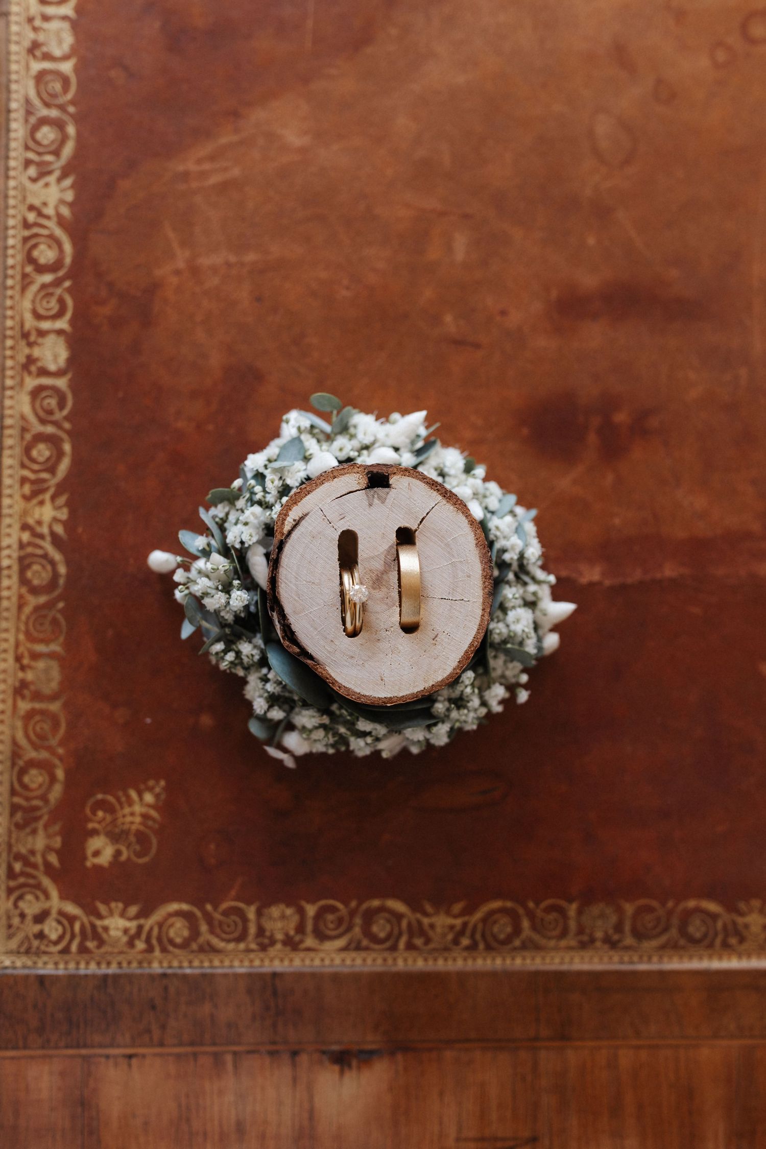 Two gold wedding bands displayed on rustic wood slice with baby's breath wreath on leather surface.