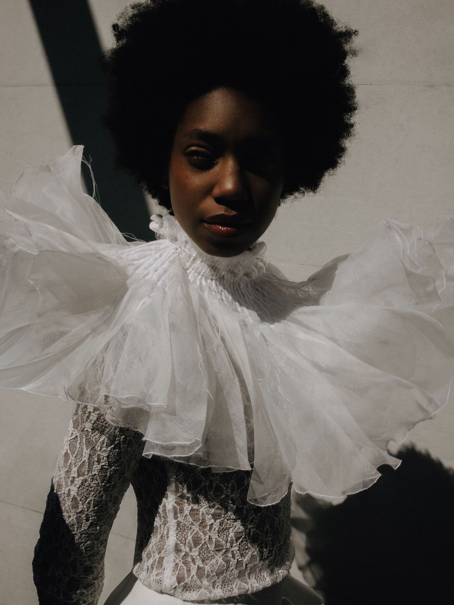 A Black woman with an afro models a white lace bodysuit with an oversized ruffled organza collar in sunlight.