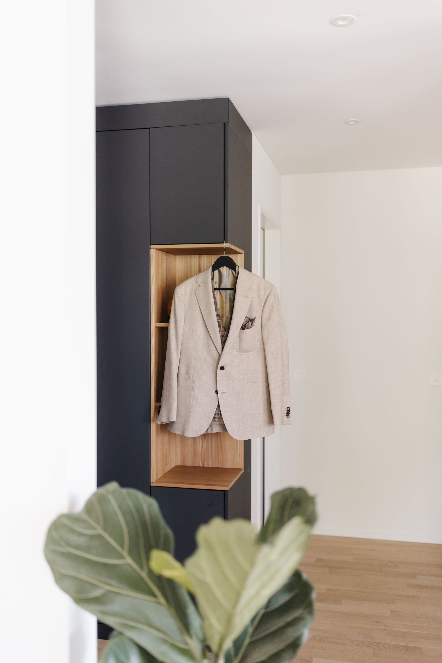 Elegant beige groom suit jacket hanging in a modern dark wardrobe with wooden shelf accents.