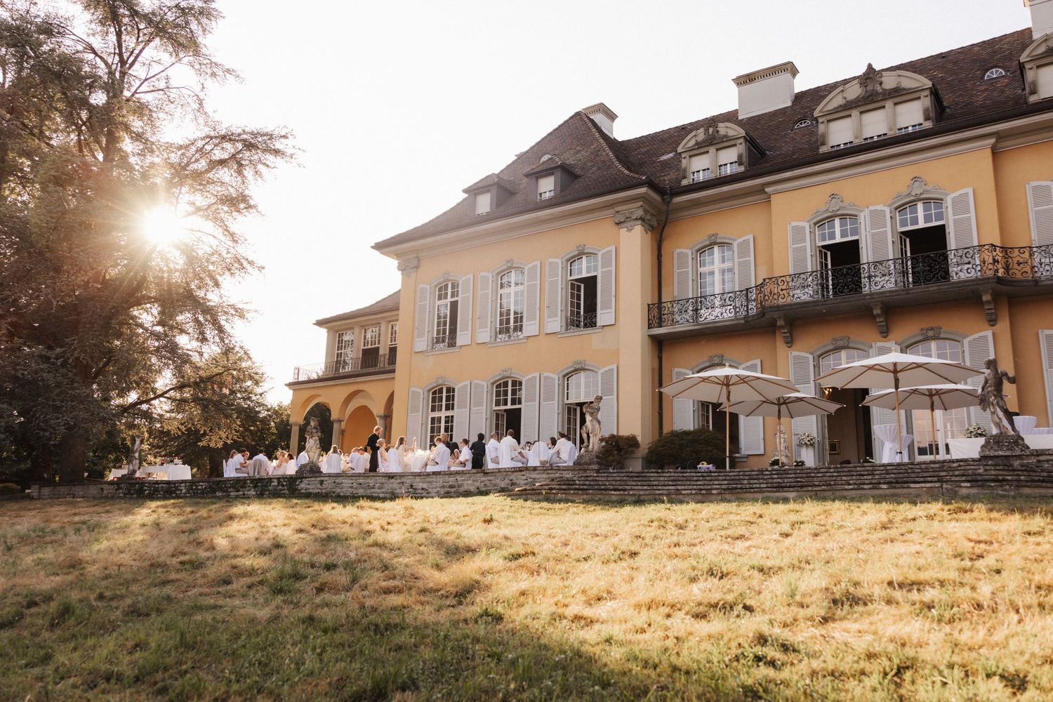 Sunlit yellow villa with wedding guests in white dining on the terrace surrounded by manicured lawns at sunset.