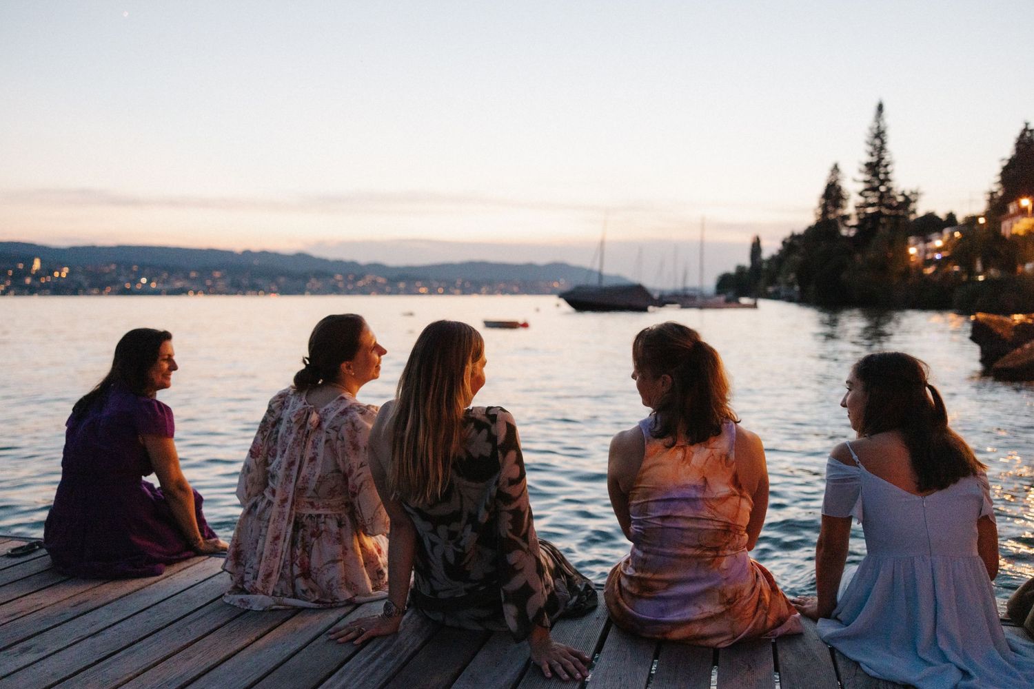 Five women sitting on a wooden dock watching a serene sunset over the water.