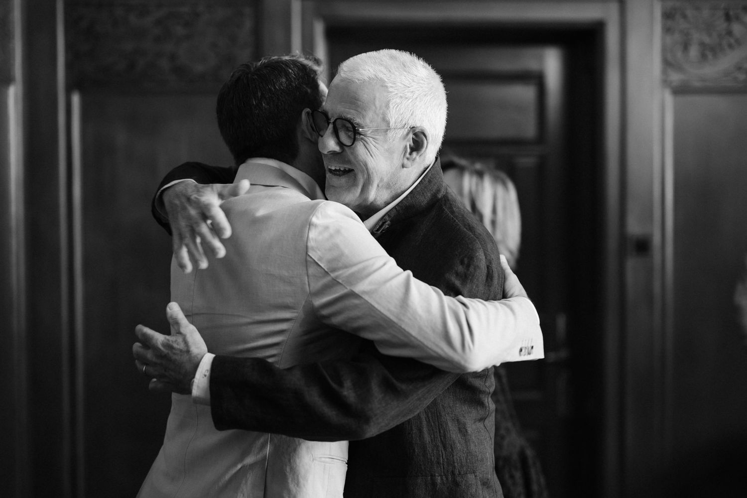 Black and white photo of groom embracing elderly man in warm congratulatory hug inside wedding venue.