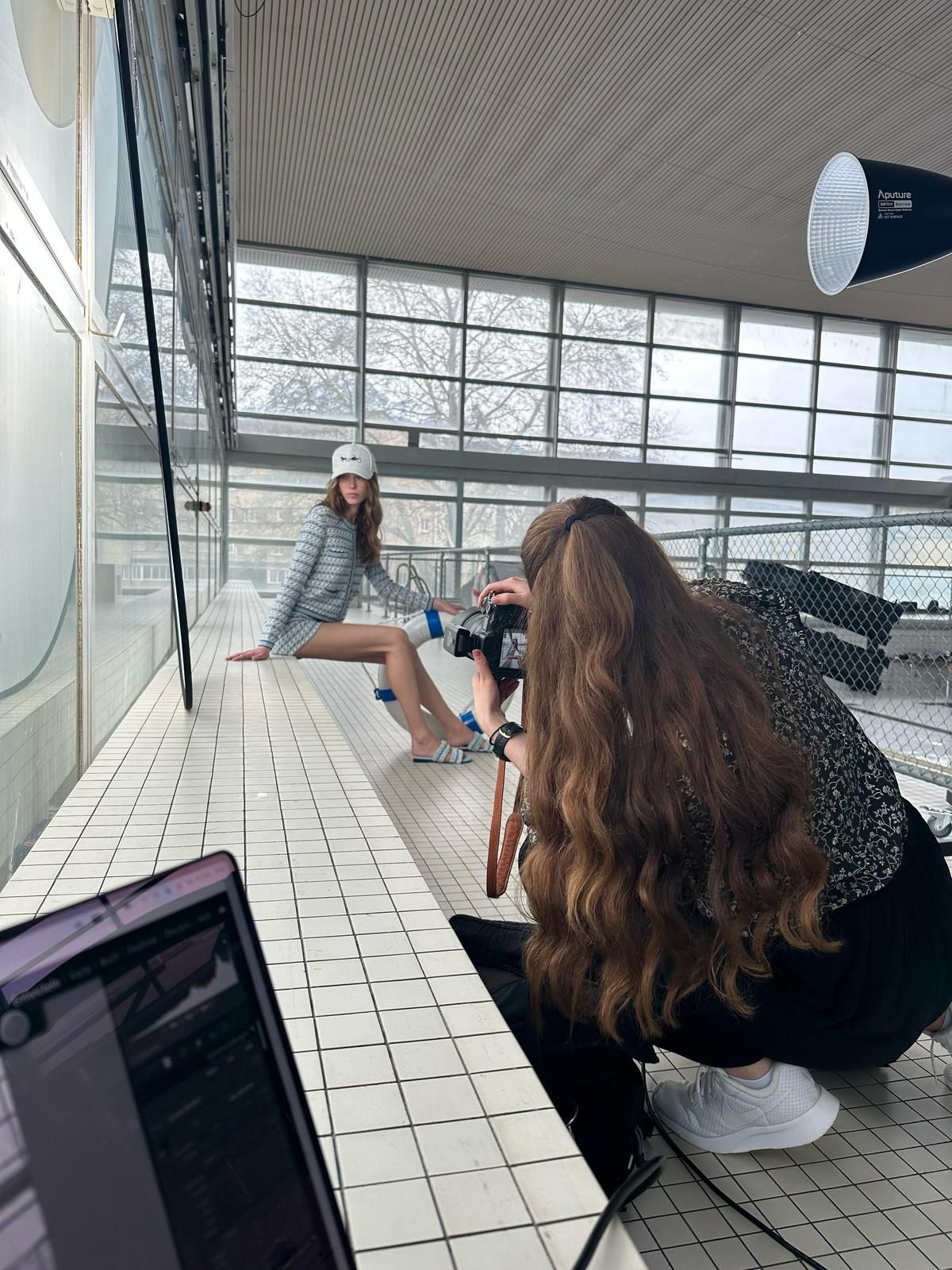 Photographer crouching to capture a seated model in striped outfit at an indoor pool setting.