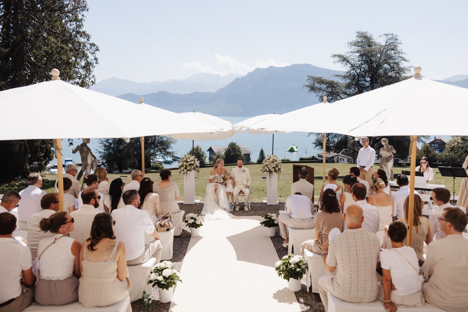 Outdoor wedding ceremony with guests seated under white umbrellas with stunning mountain and lake backdrop.