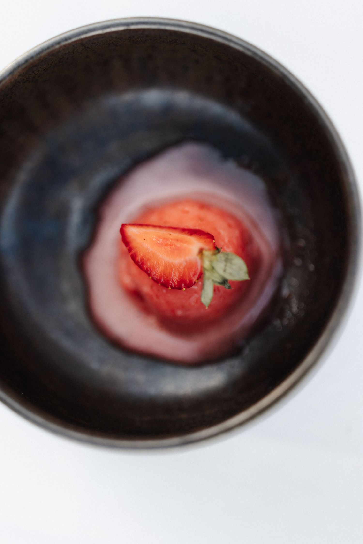 Overhead view of a fresh sliced strawberry in a dark ceramic bowl with pink sauce.