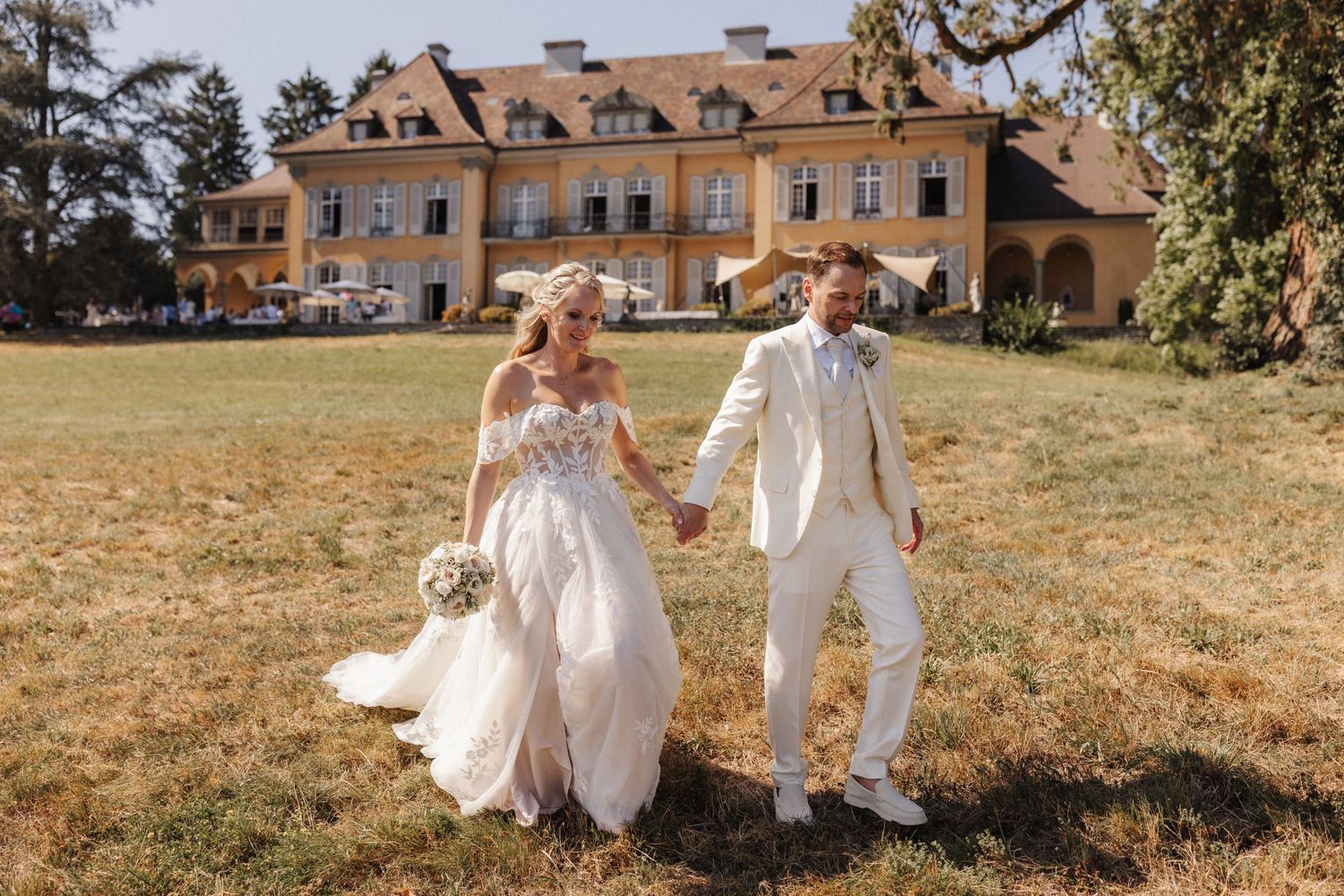 Bride and groom walking hand in hand across a sunlit lawn in front of a grand chateau.
