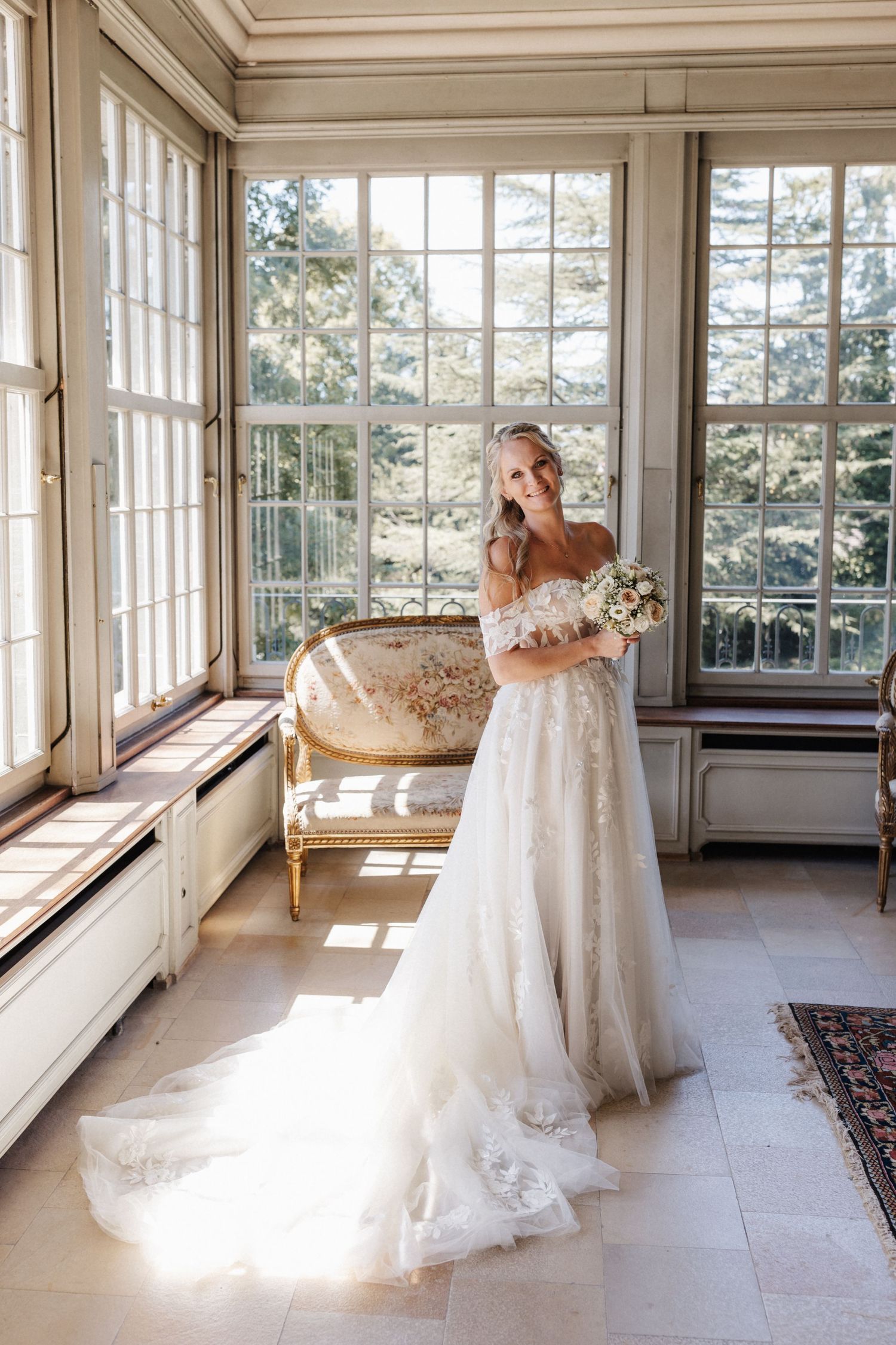 Bride in off-shoulder lace wedding gown holding bouquet in sunlit elegant room with tall windows.