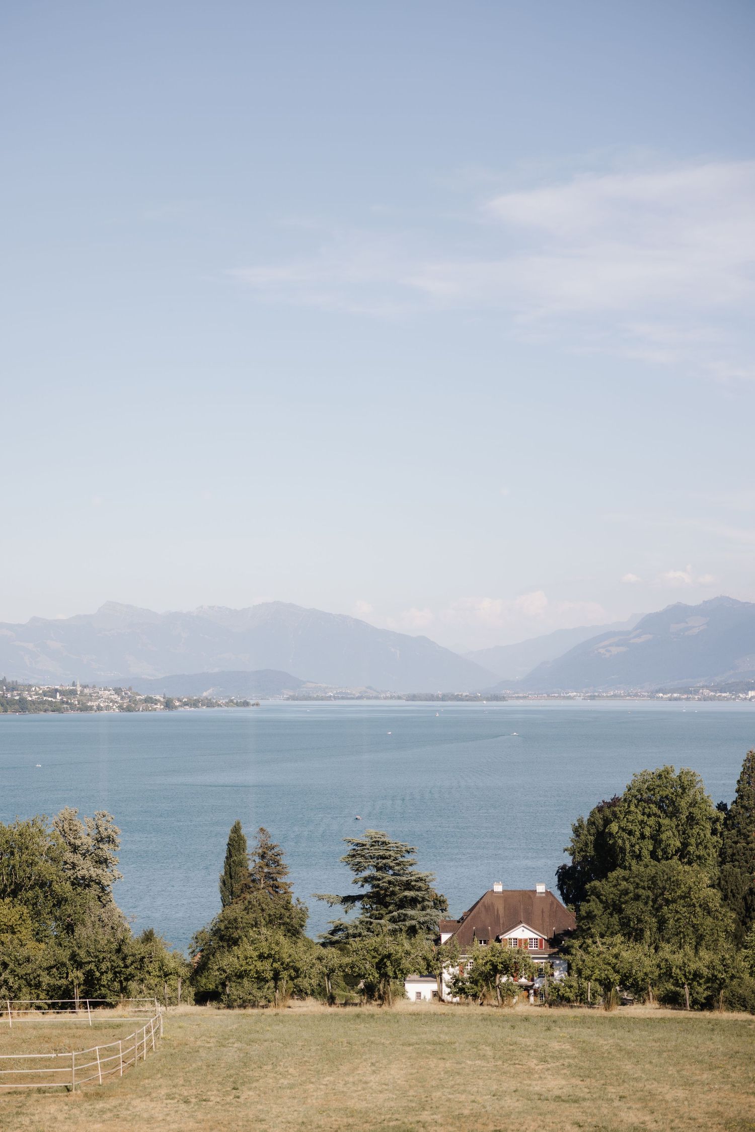Scenic lakeside view with mountains in background, green trees, grassy field, and a house near the shore.