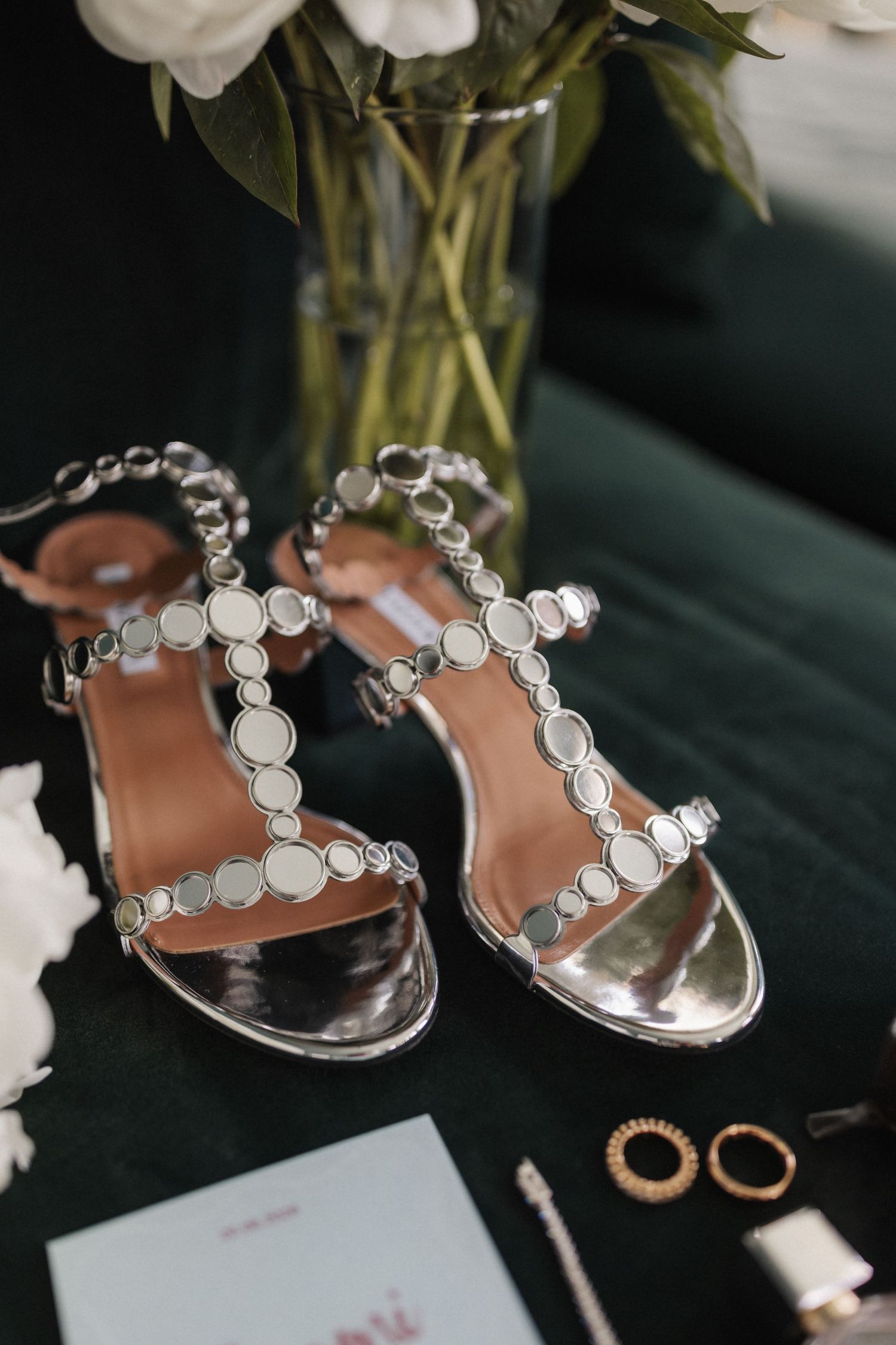 Silver embellished bridal heels displayed on green velvet beside white flowers.