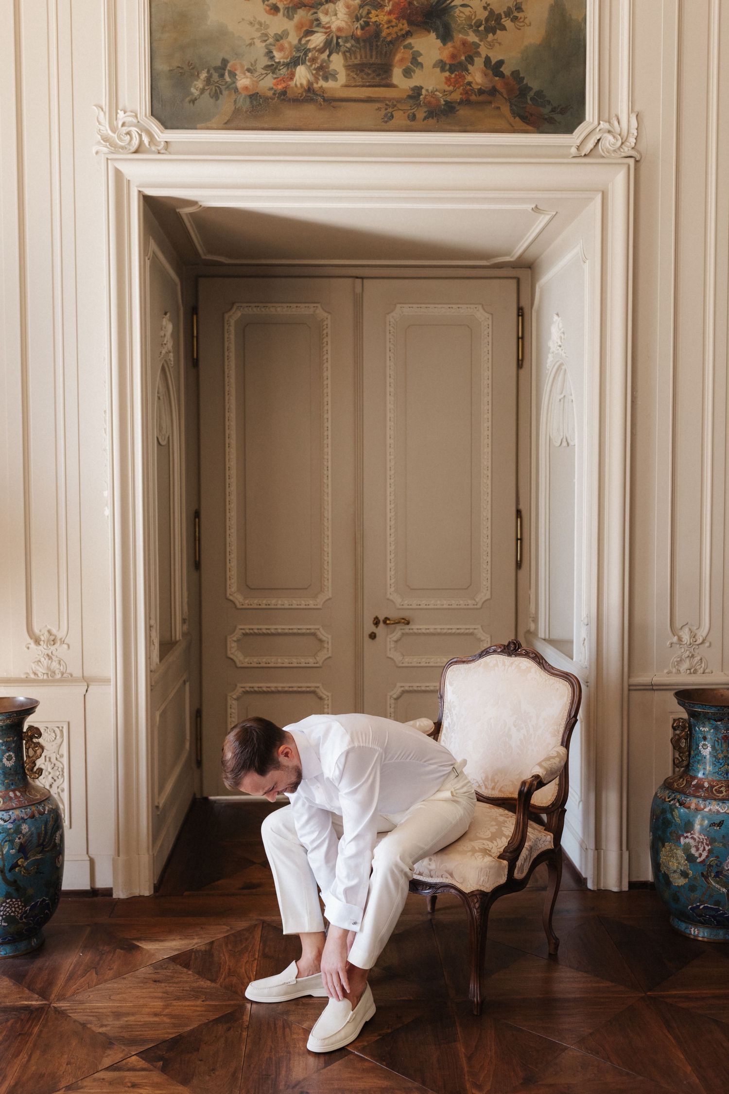Groom in white suit putting on shoes in elegant French chateau room with ornate moldings.