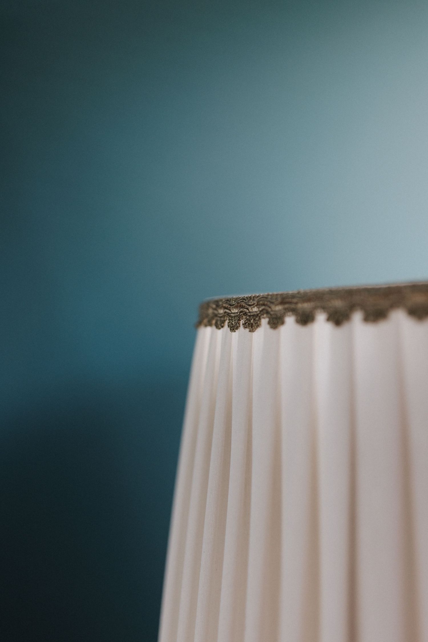 Close-up of a pleated white lamp shade with gold trim against a teal background.
