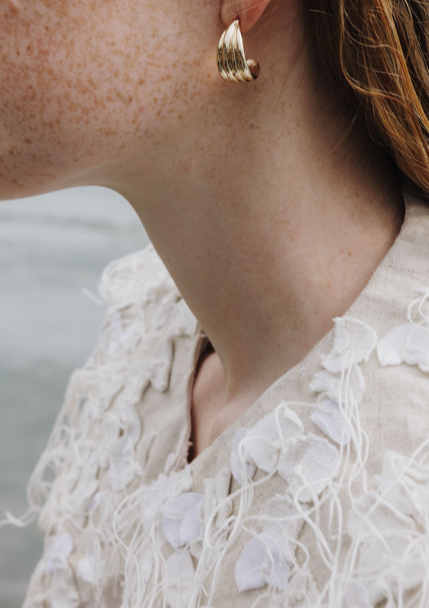 Close-up of a freckled woman wearing a gold sculptural earring and white textured top.