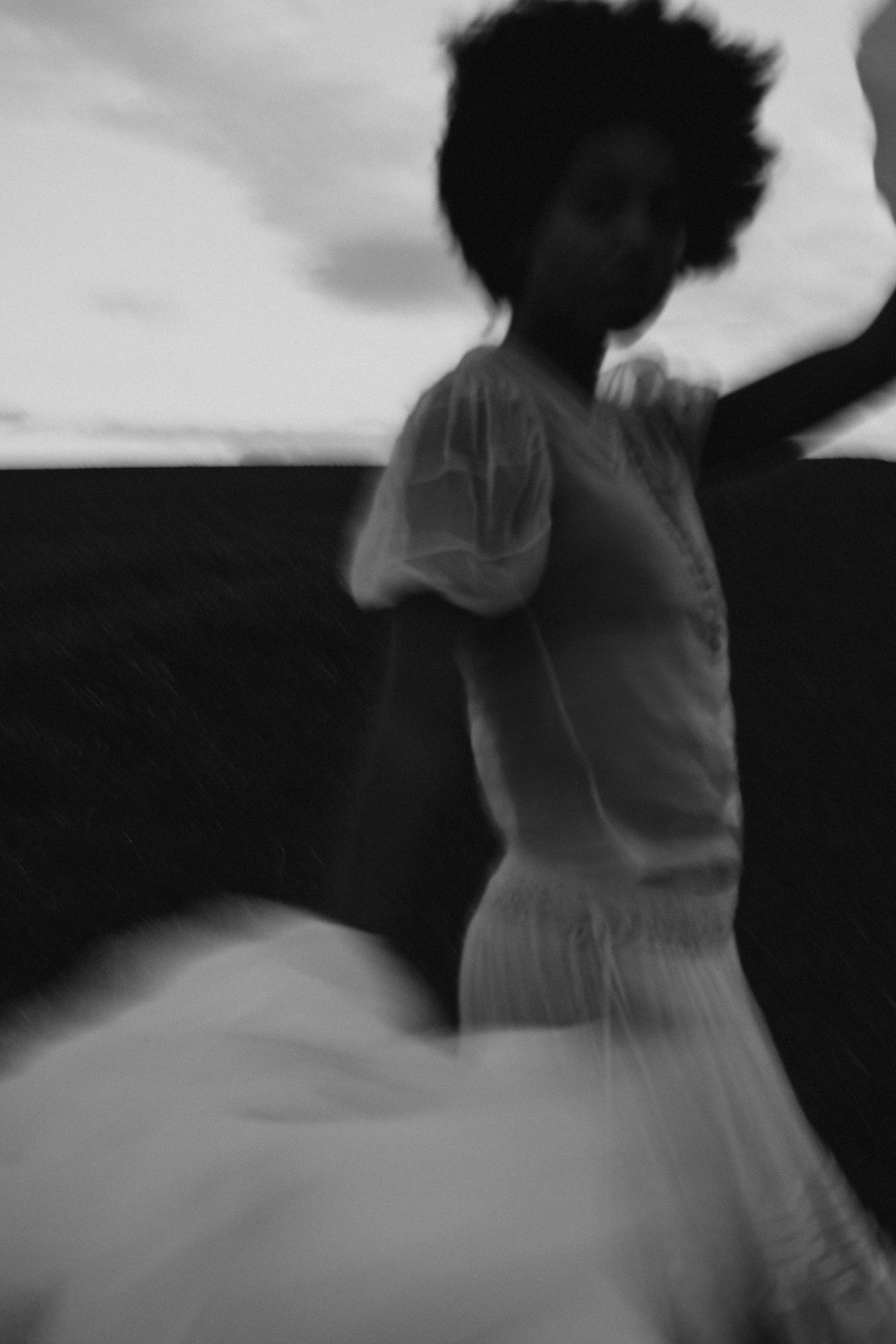 Blurred black and white photo of a woman with an afro dancing in a white dress outdoors.