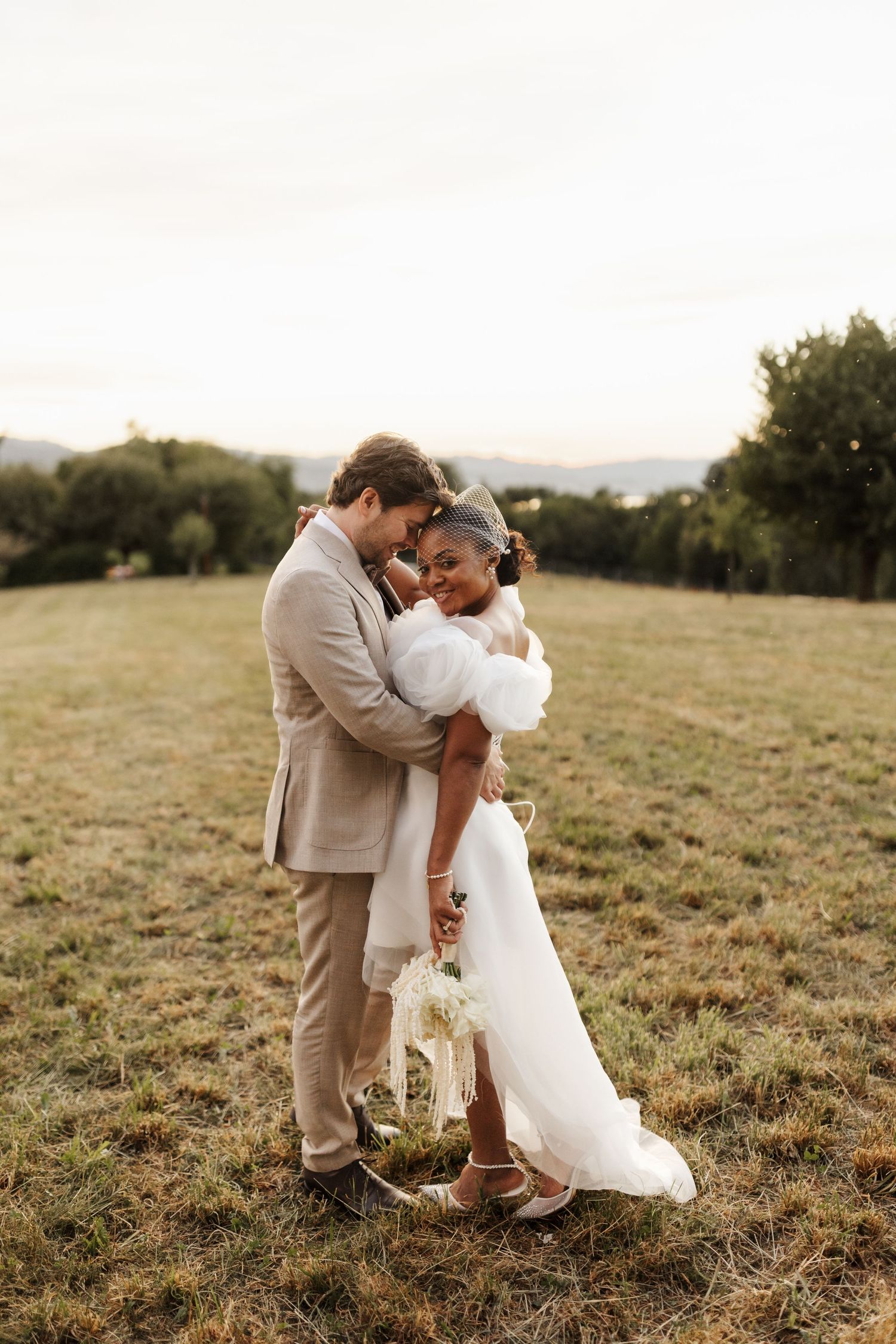 Interracial couple embracing in a field at golden hour, bride in white gown with bouquet, groom in beige suit.
