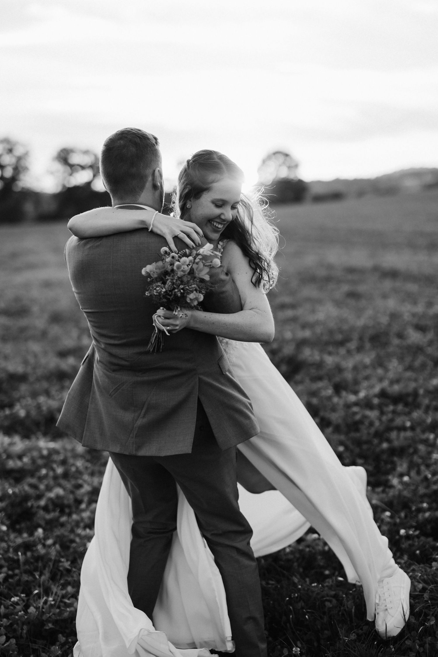 Black and white photo of groom lifting laughing bride in a sunlit field.