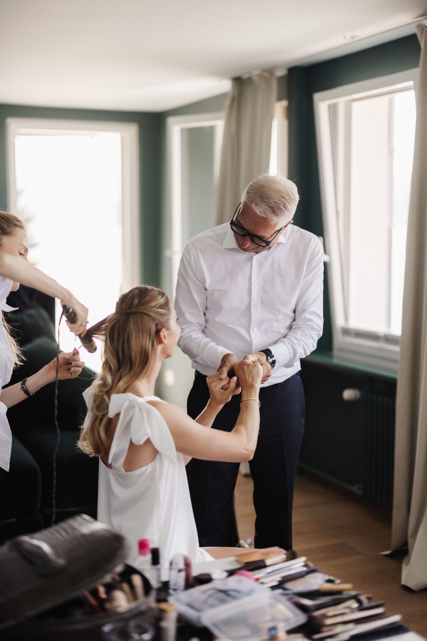 Older man holding bride's hands while hairstylist works in bright room.