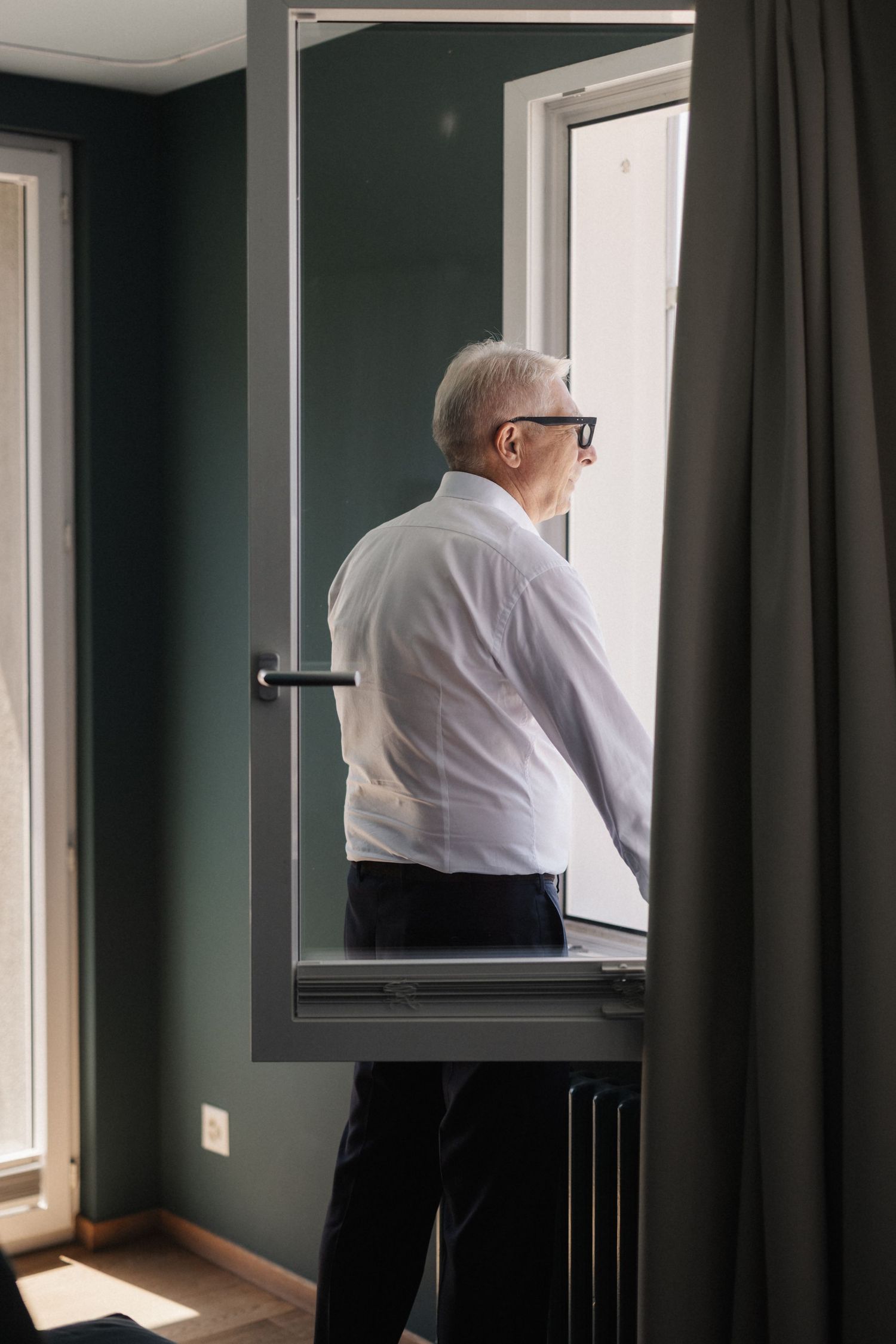 Elderly man in white shirt and glasses looking out an open window in a dark green room.