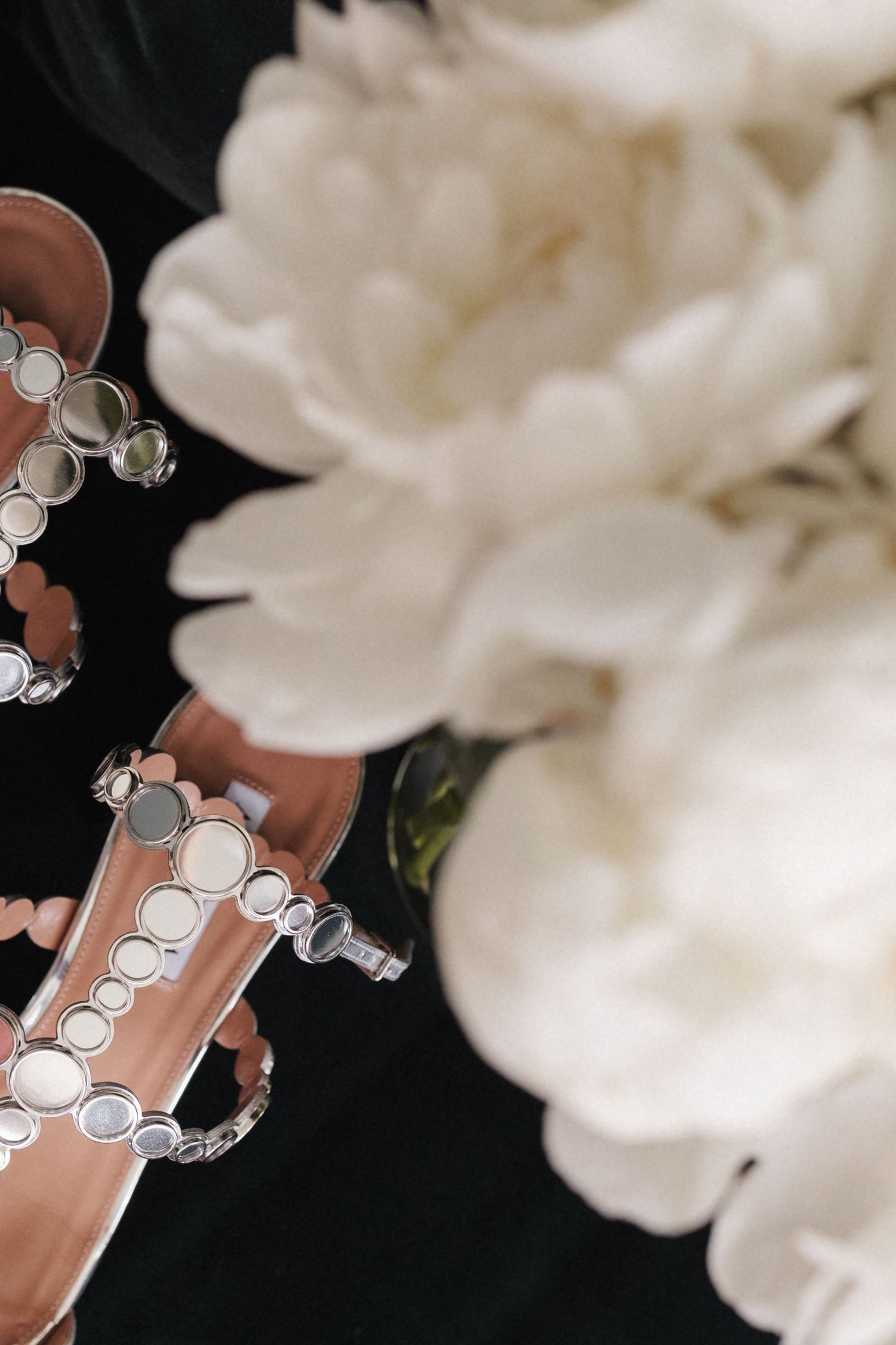 Rose gold studded bridal sandals beside white peony flowers on a dark background.