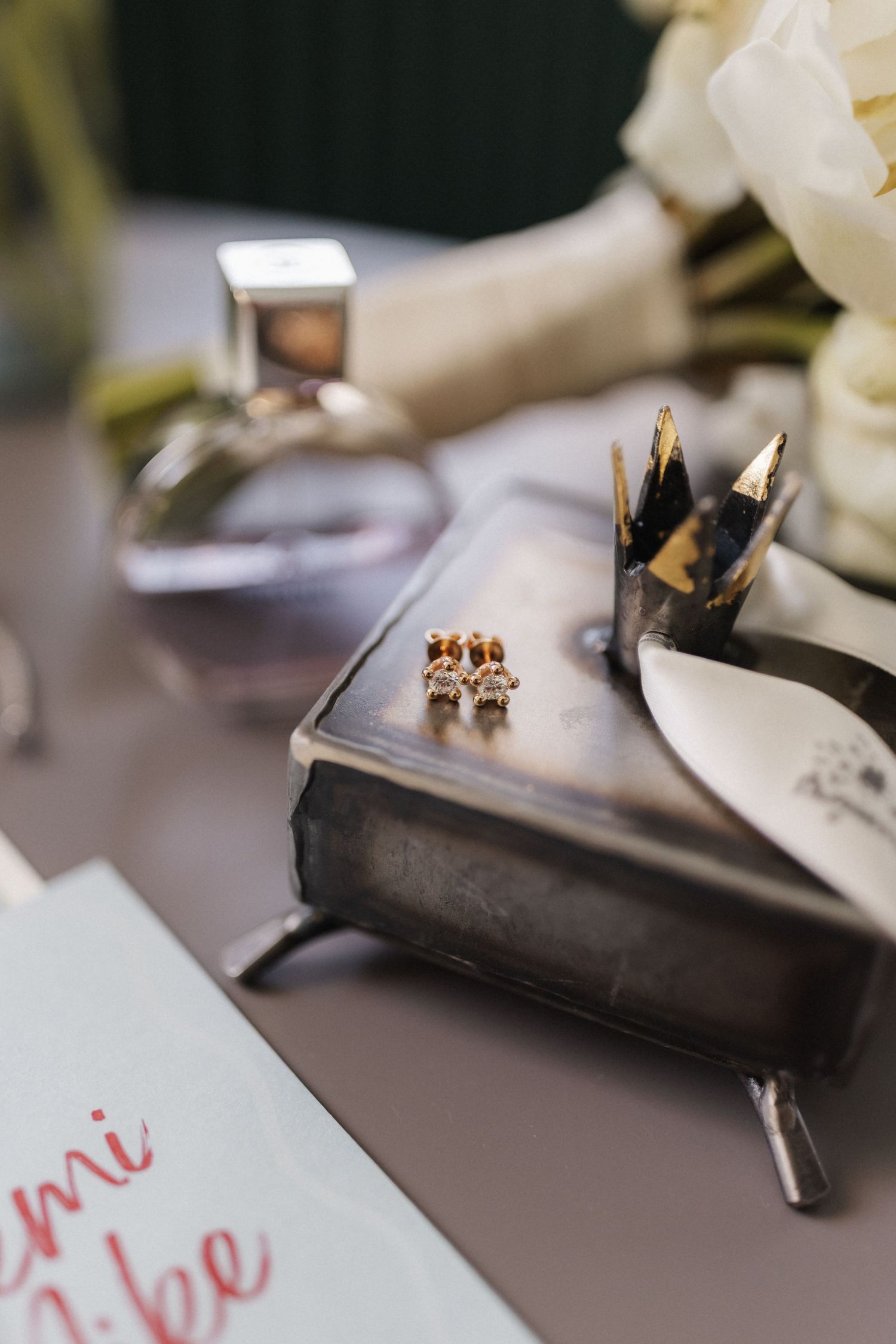 Gold diamond stud earrings placed on a vintage silver jewelry box near a white floral bouquet.