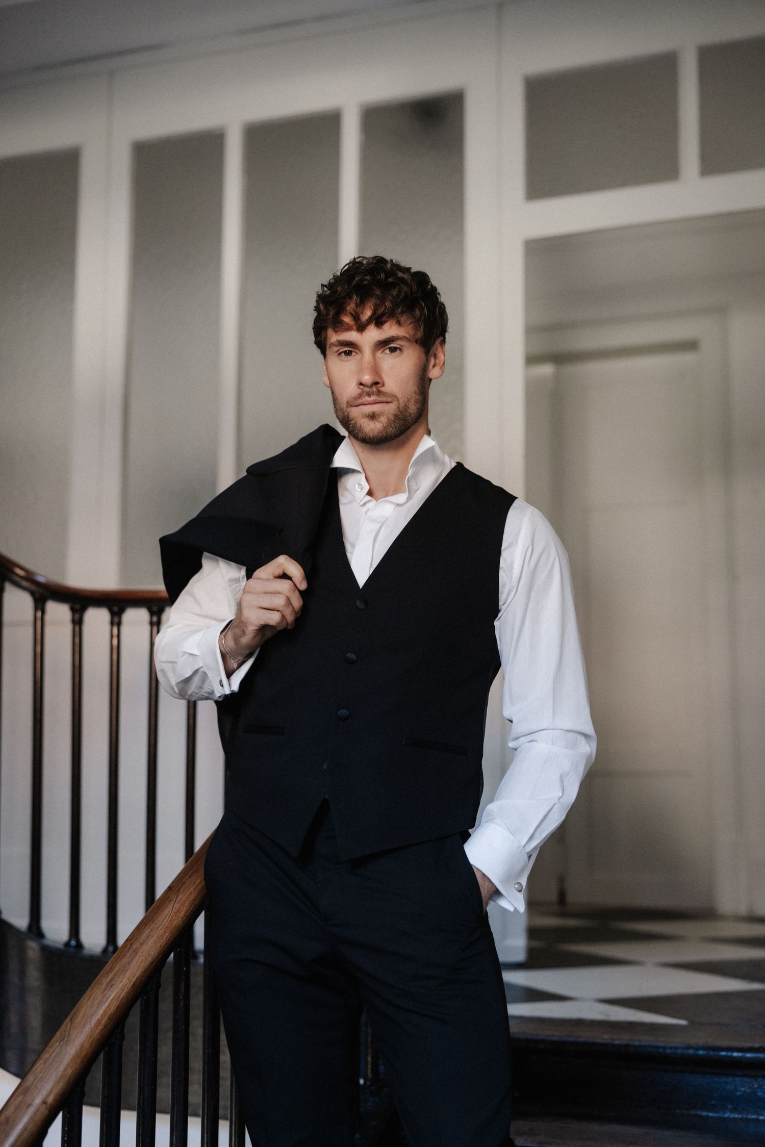 Handsome groom in black vest and white shirt holding jacket over shoulder near staircase railing.