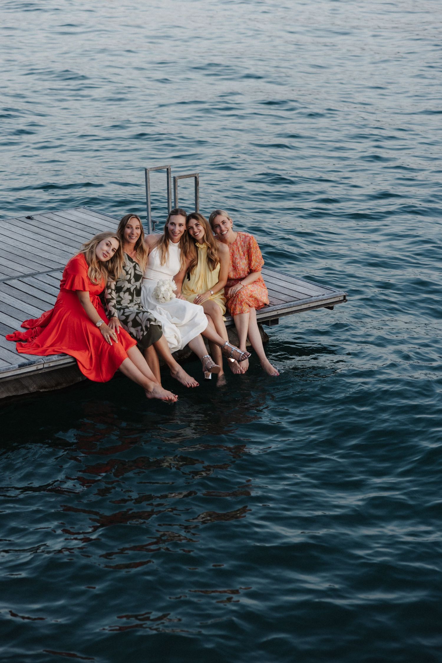 Bride and four friends in colorful dresses sit barefoot on a wooden dock edge over blue water.