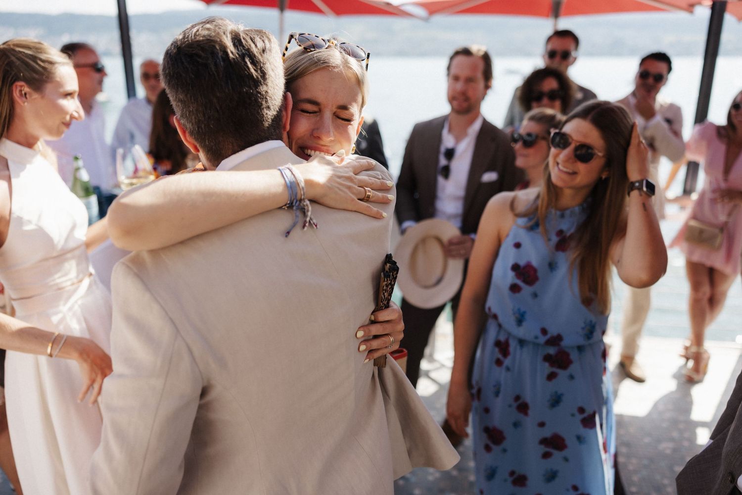 Wedding guests sharing warm embraces at a sunny outdoor waterfront celebration.
