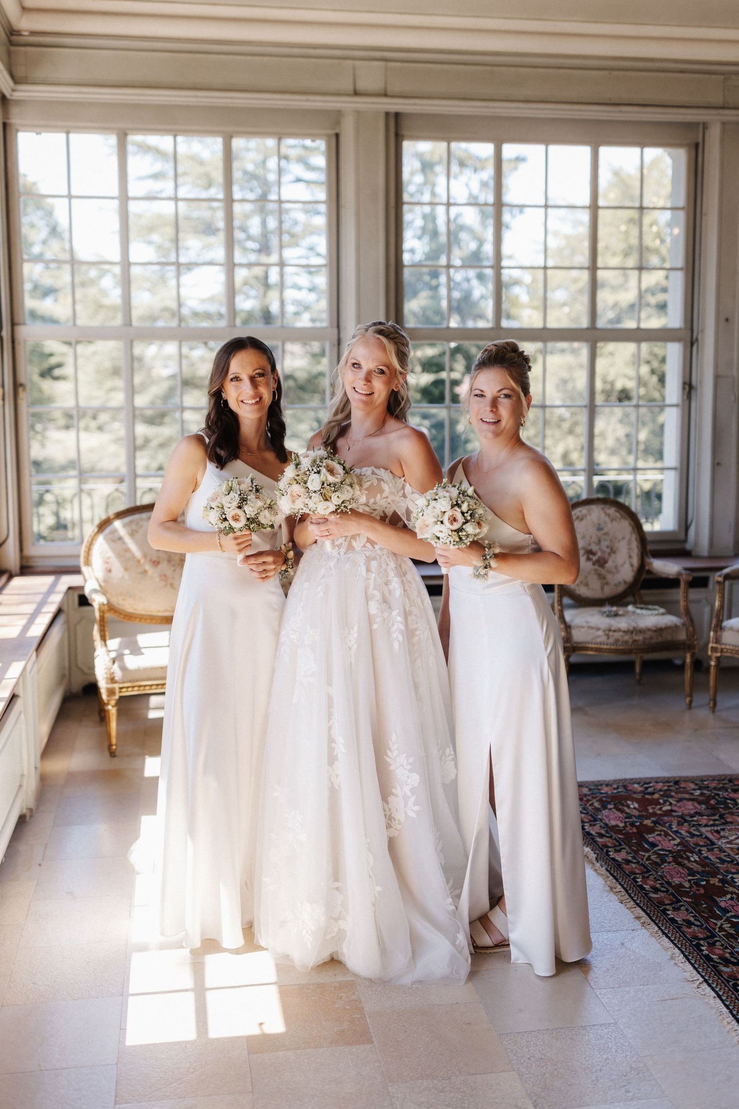 Bride in a floral lace wedding gown poses with two bridesmaids in white dresses holding bouquets indoors.
