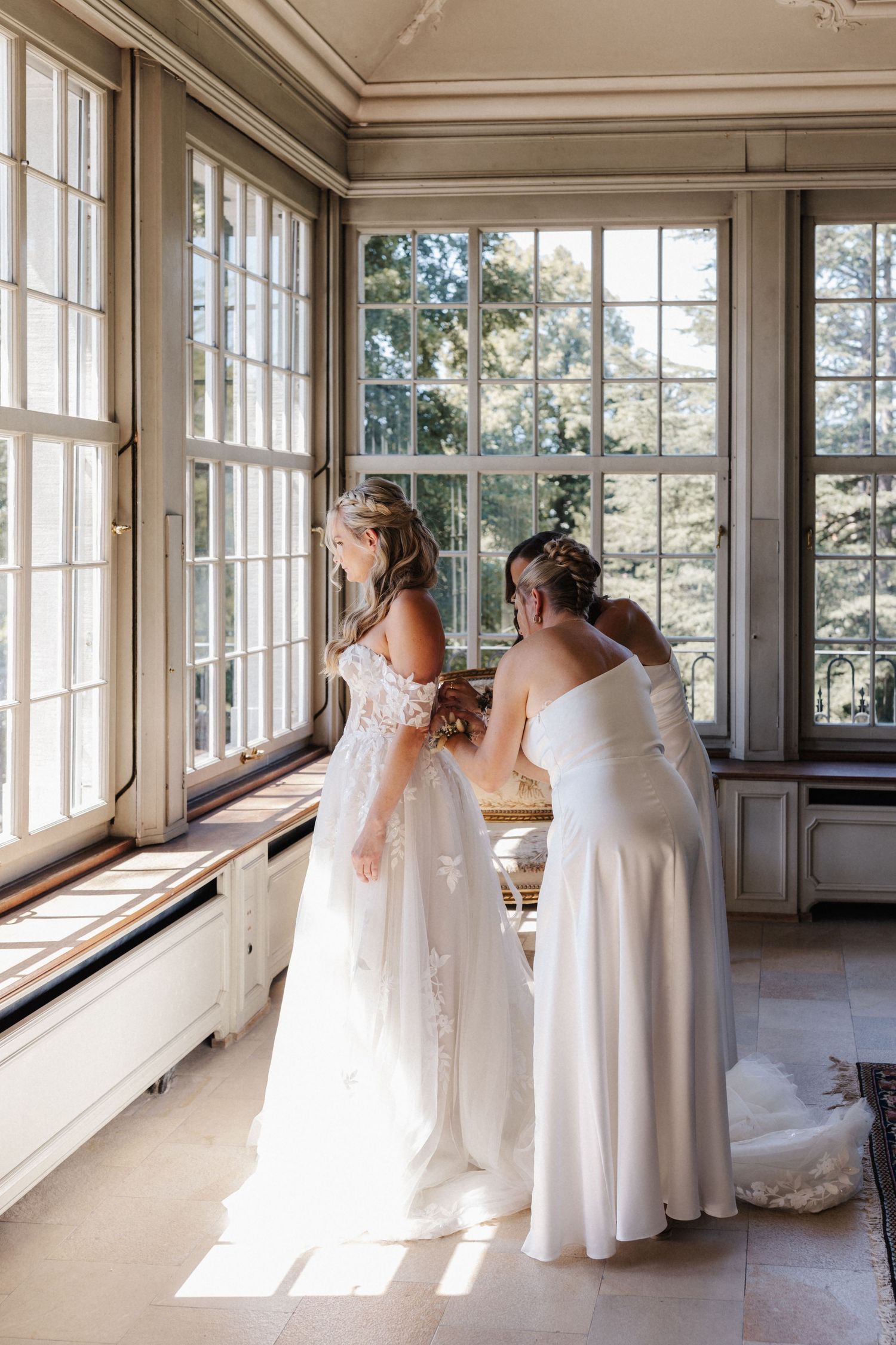 Two brides in white gowns help each other get ready by large windows in an elegant sunlit room.