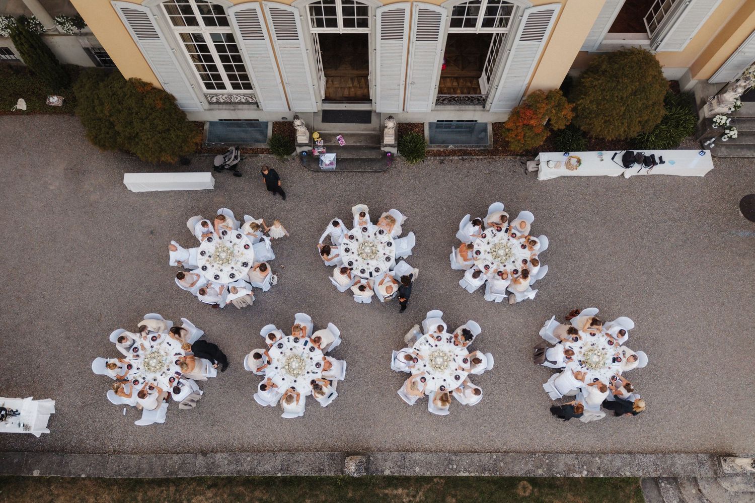 Aerial view of an outdoor wedding reception with round white-draped tables arranged outside a yellow manor house.