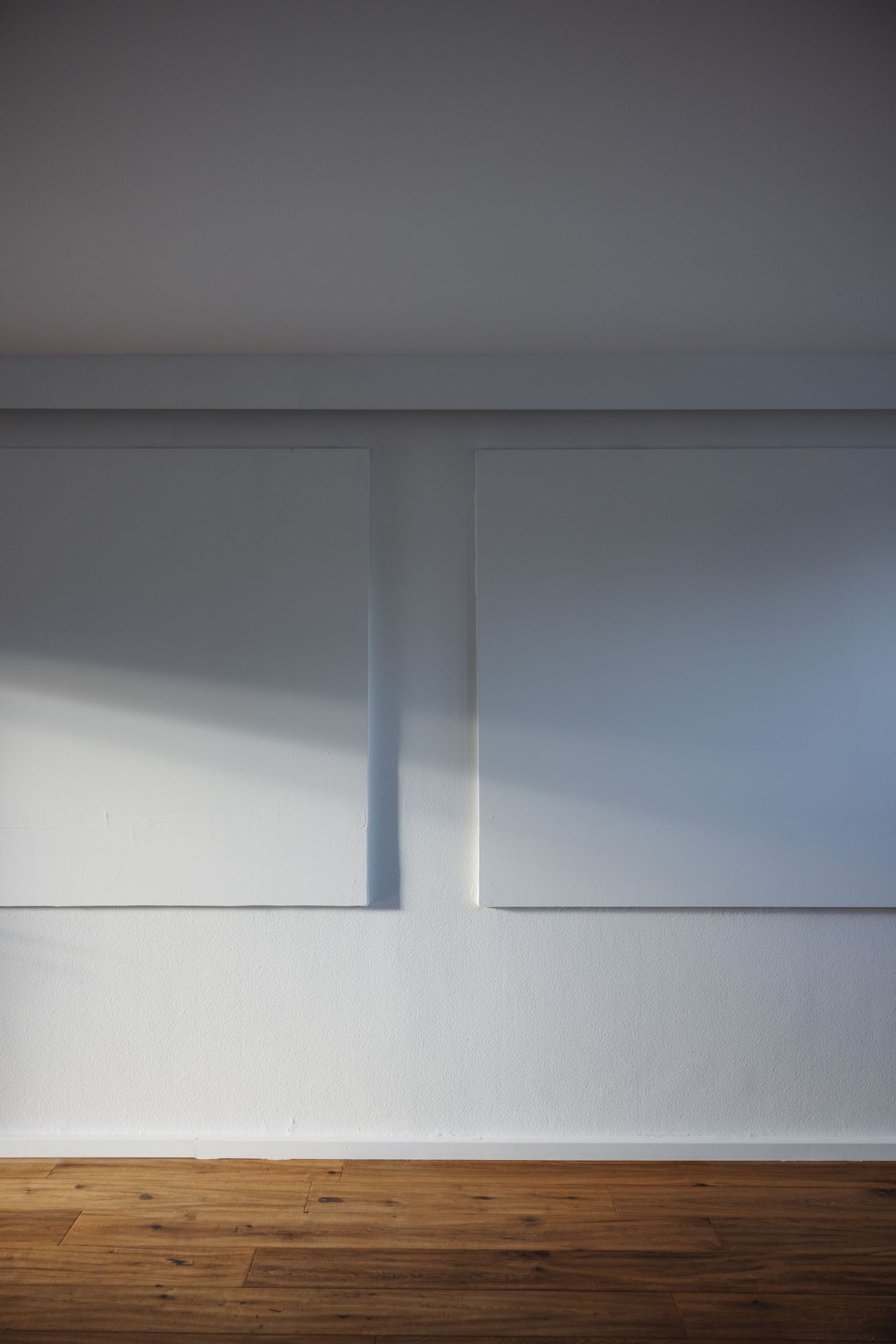 Two blank white canvases hang on a white gallery wall above a warm wooden plank floor, lit by soft natural light and shadow.