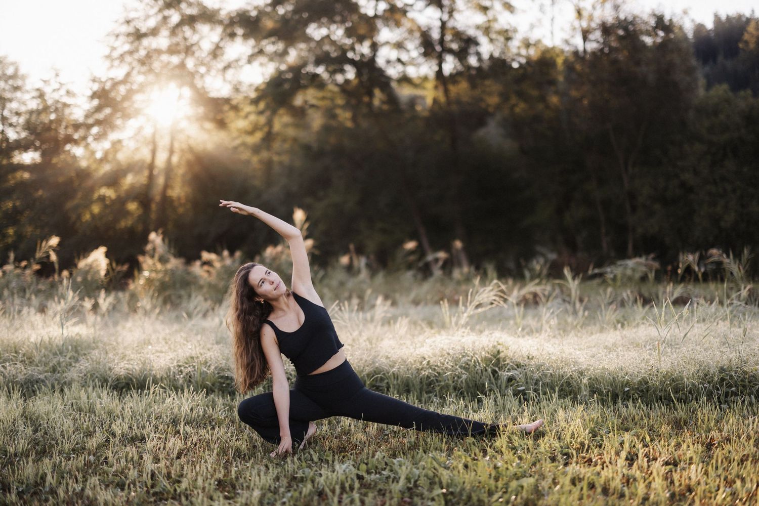 Woman in black activewear doing yoga pose in a sunlit meadow at sunset.