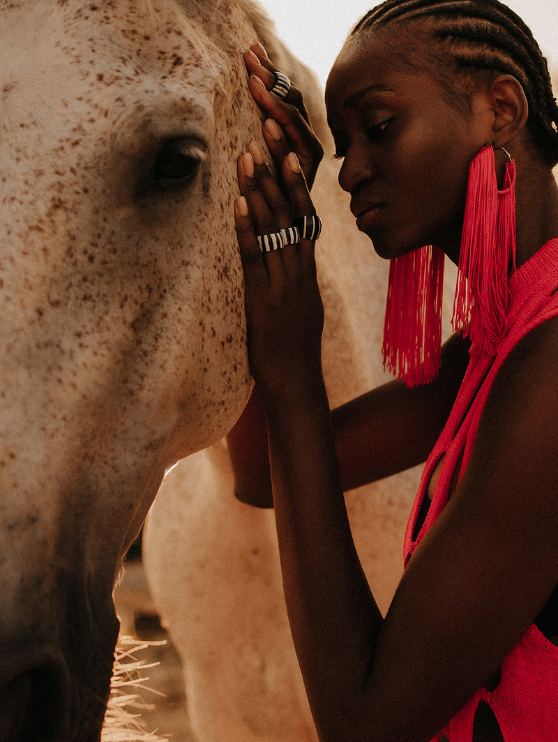 Woman in red dress with fringe earrings tenderly touching a white horse face in warm light.