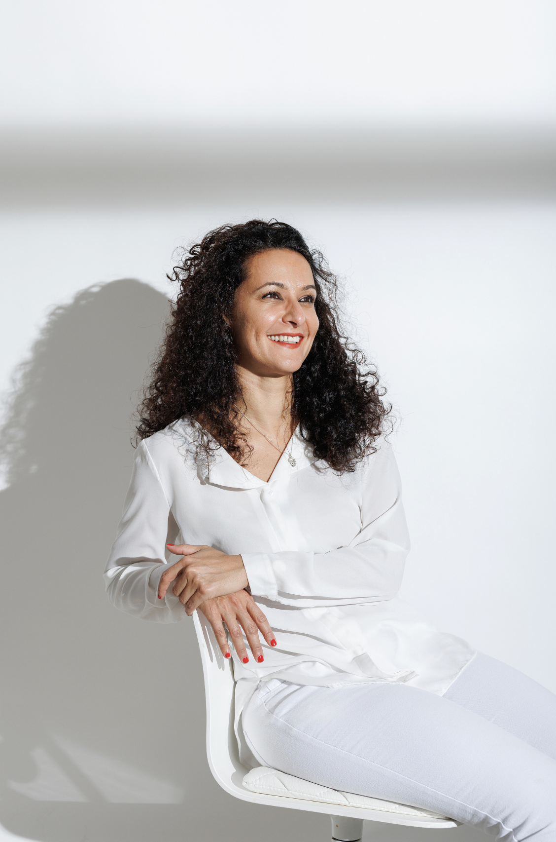 Smiling woman with curly hair in white outfit sitting on chair against white background.