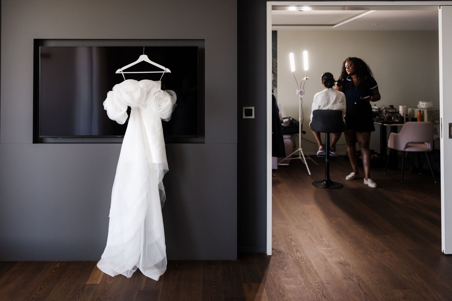White wedding dress with puffy sleeves hanging on wall while bride gets ready in background.