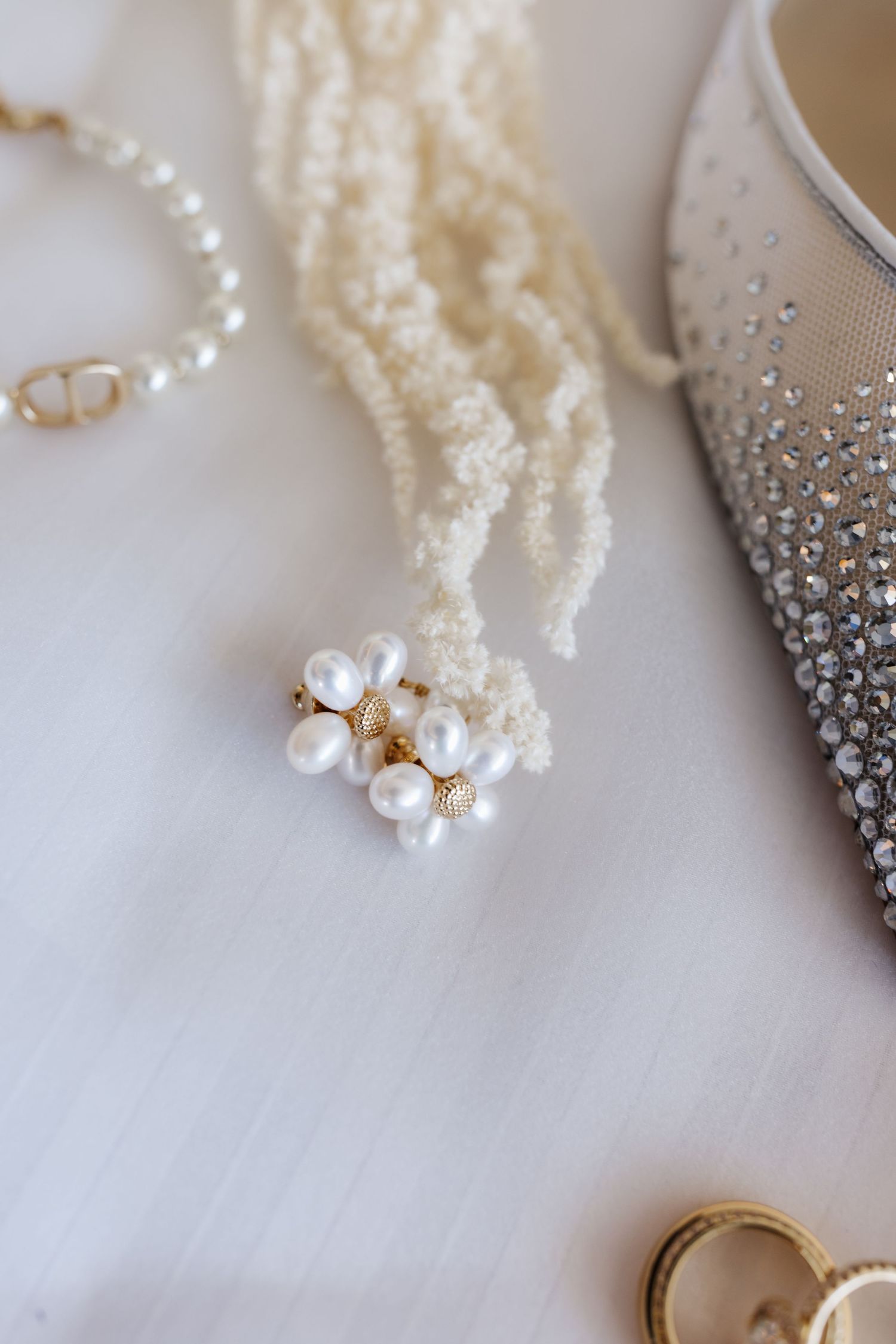 Pearl cluster earrings resting on ivory beaded necklace beside crystal bridal shoe on white fabric.