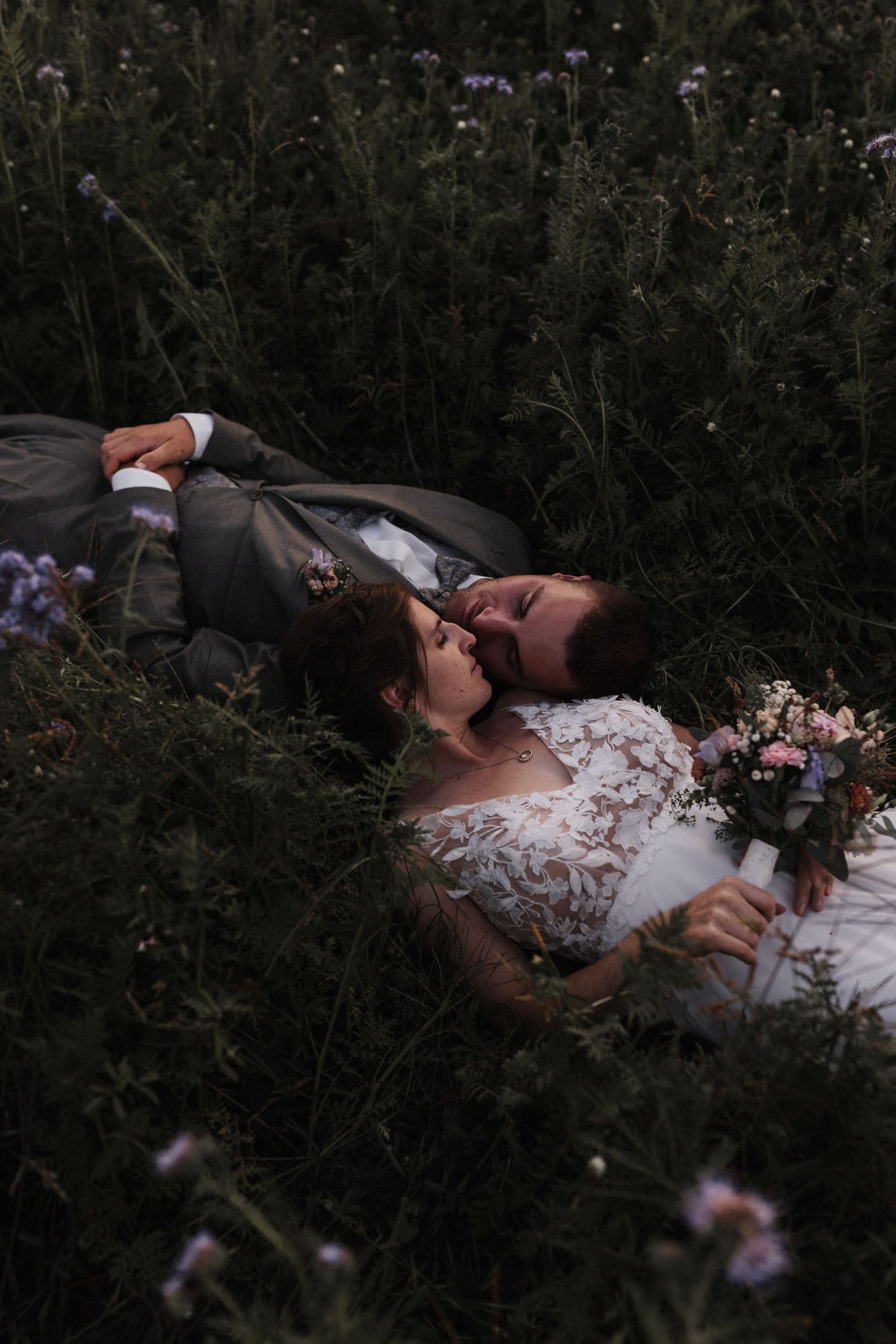 Bride and groom laugh together while lying in a lush wildflower field holding a bridal bouquet.