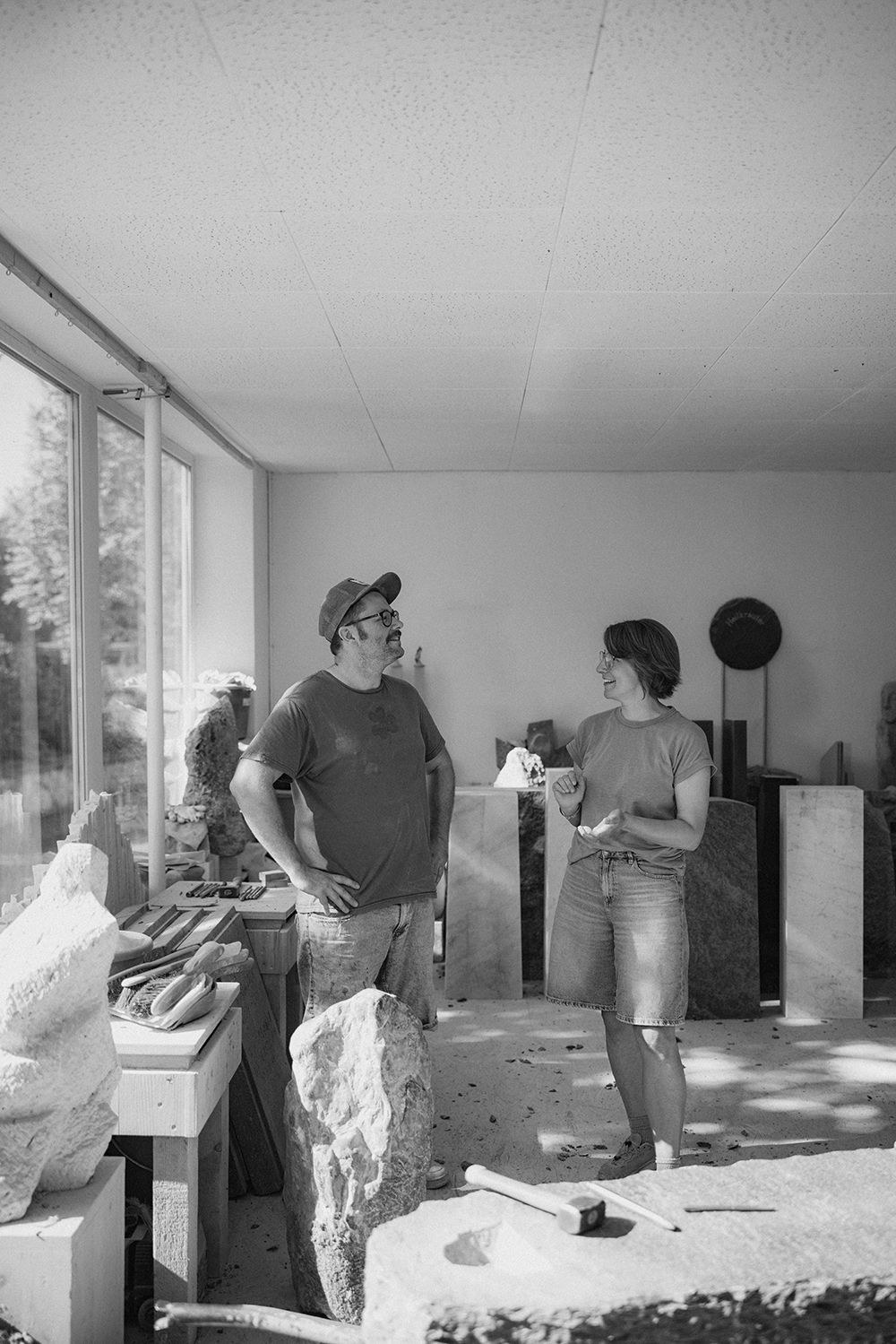 Two sculptors discuss their work surrounded by stone sculptures and tools in a bright studio workspace.
