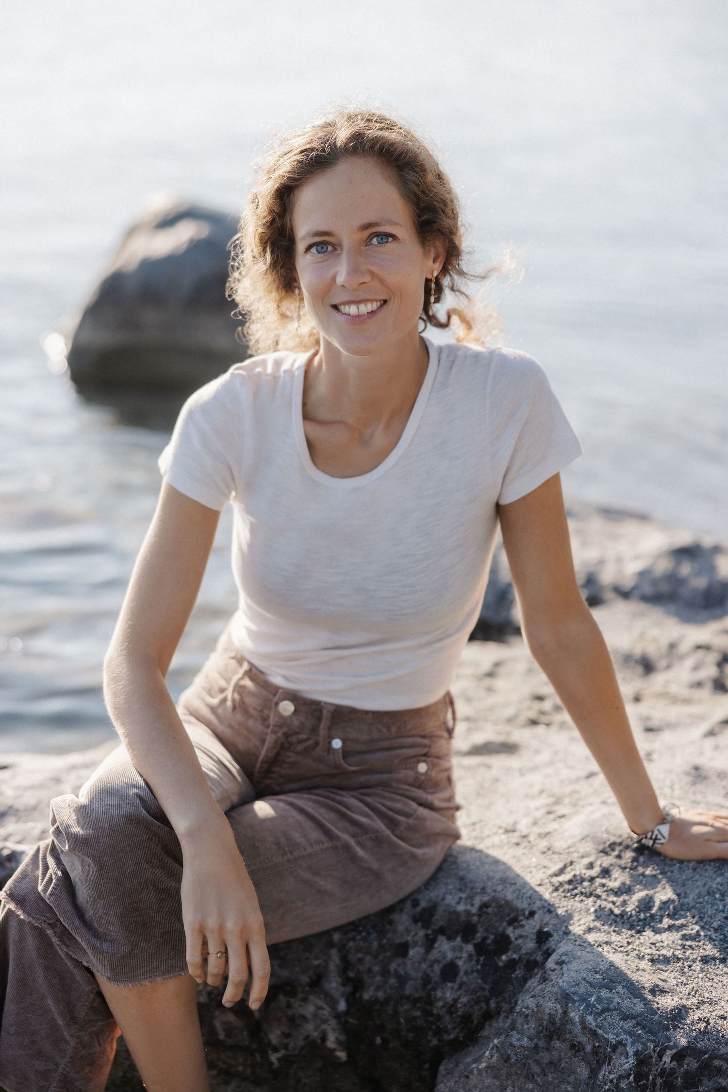 Smiling woman in white t-shirt and brown pants sitting on rocks by a sparkling lake or sea.