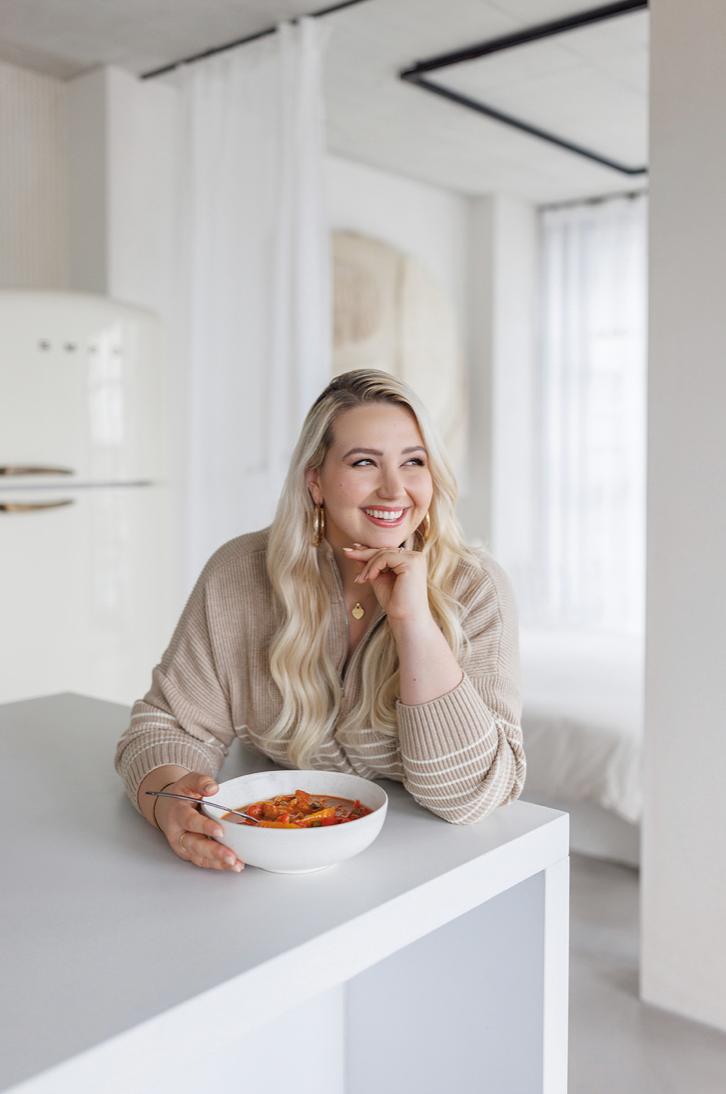 Smiling blonde woman in beige sweater holding a bowl of pasta at a white kitchen counter.