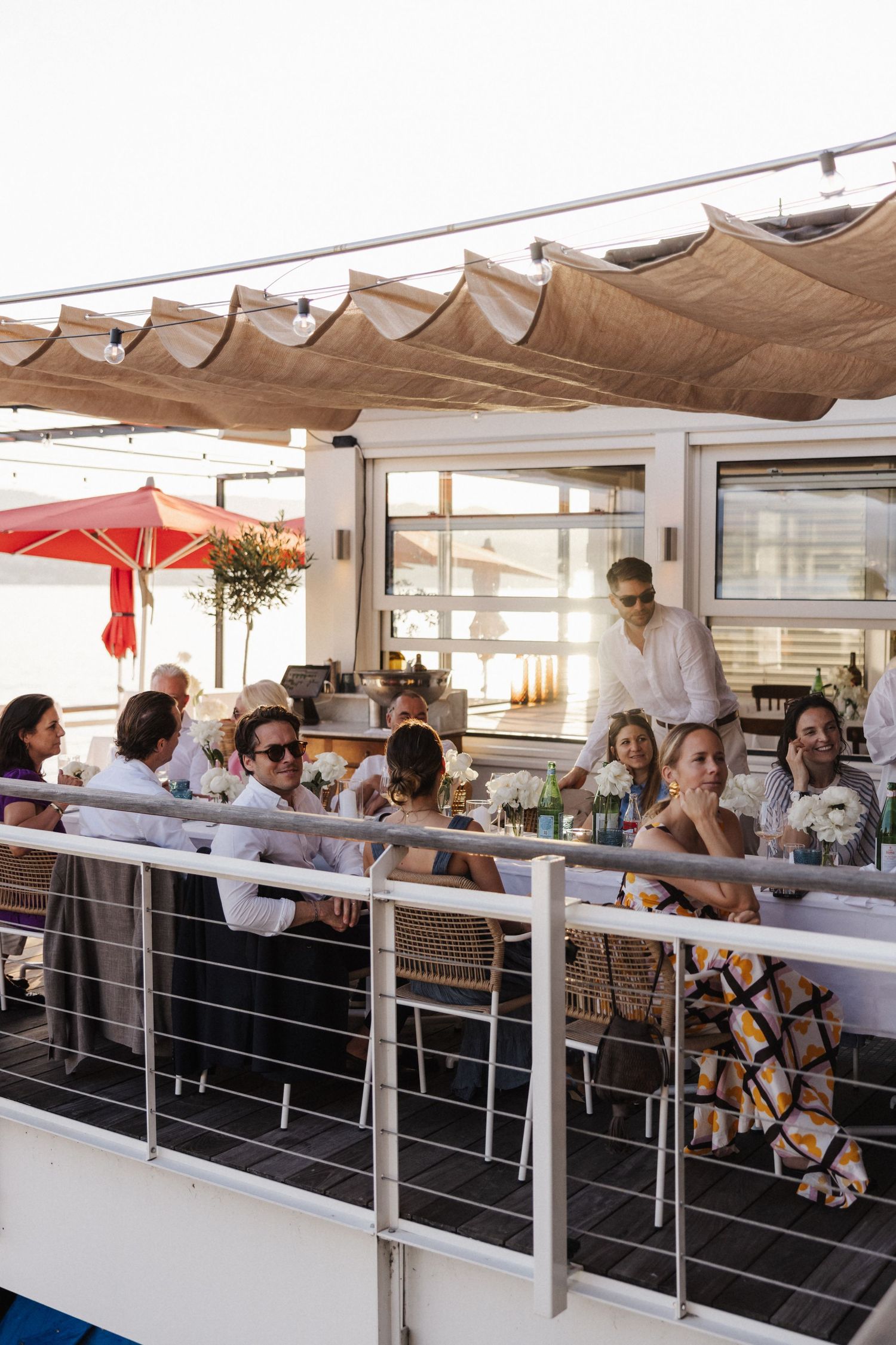 Wedding guests seated at an outdoor waterfront reception venue under a canopy at sunset.