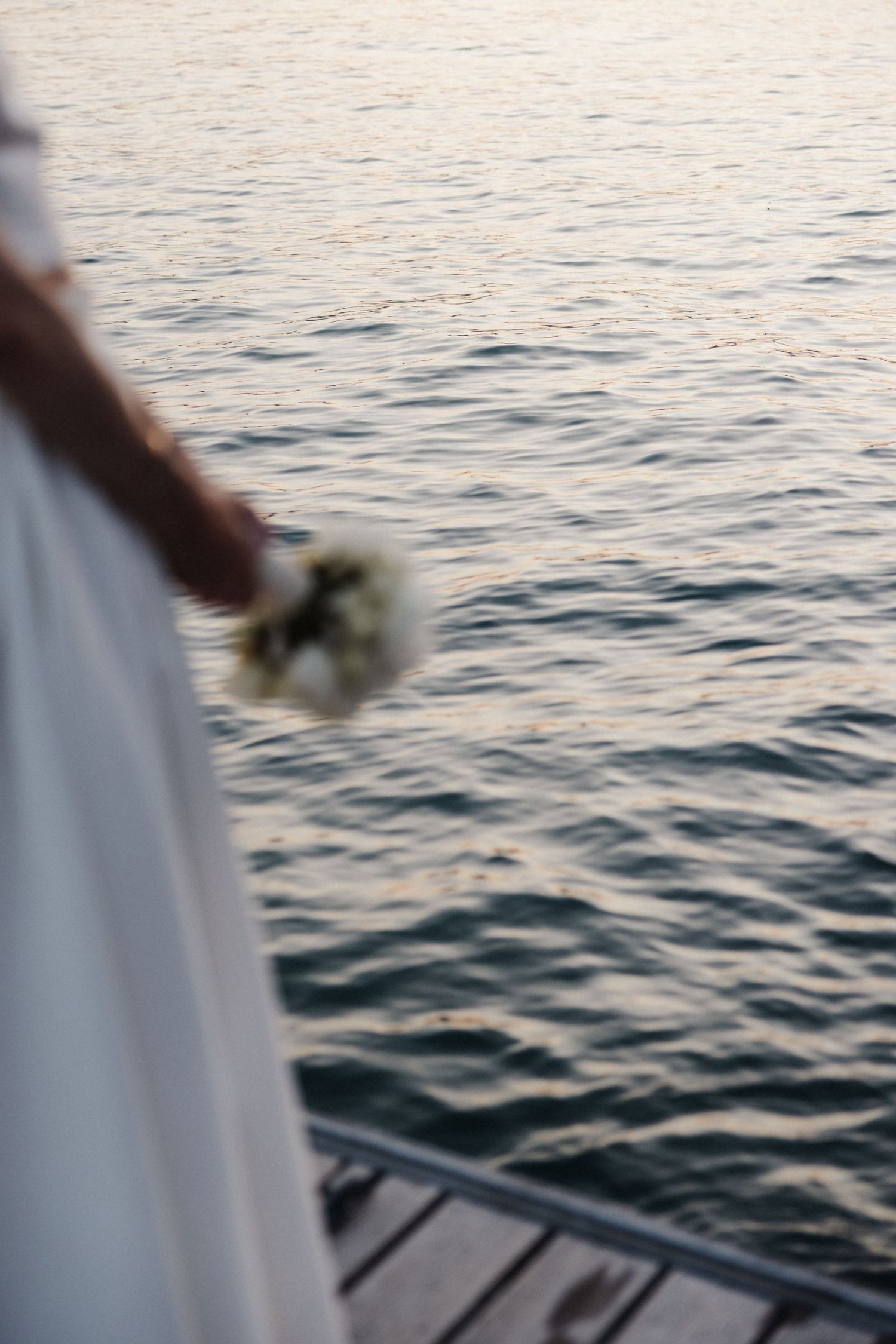 Bride in white gown holds a white bouquet while standing on a dock overlooking calm water.
